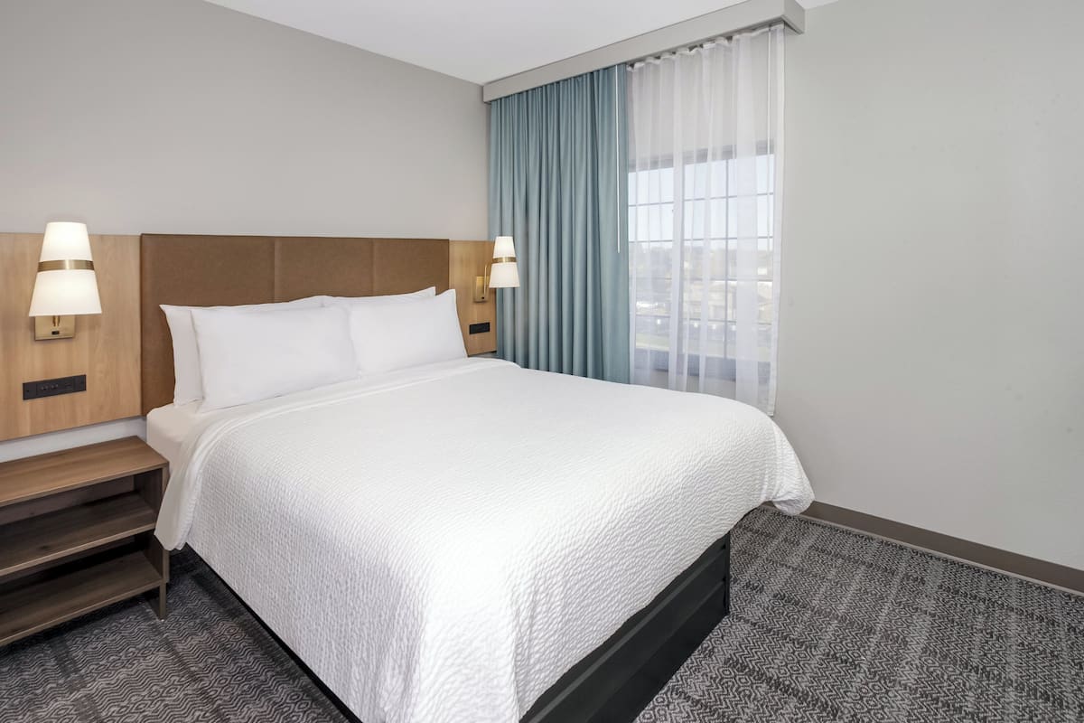 Hotellihuone, jossa on suuri sänky, kaksi yöpöytää valaisimineen, ikkuna verhoineen ja kuviollinen matto.