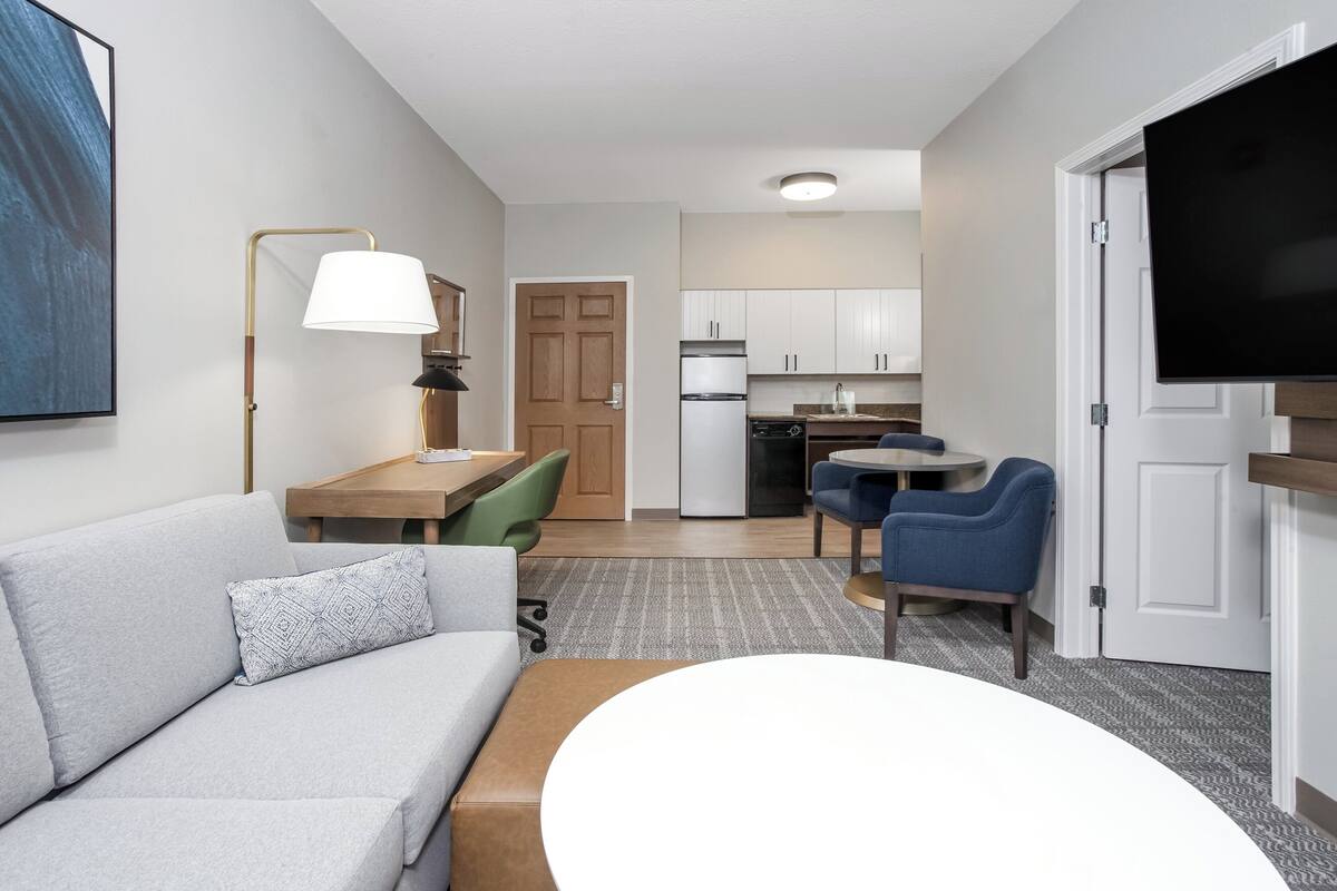 Moderni hotellihuone, jossa on sohva, ruokapöytä, televisio ja pieni keittiönurkkaus.