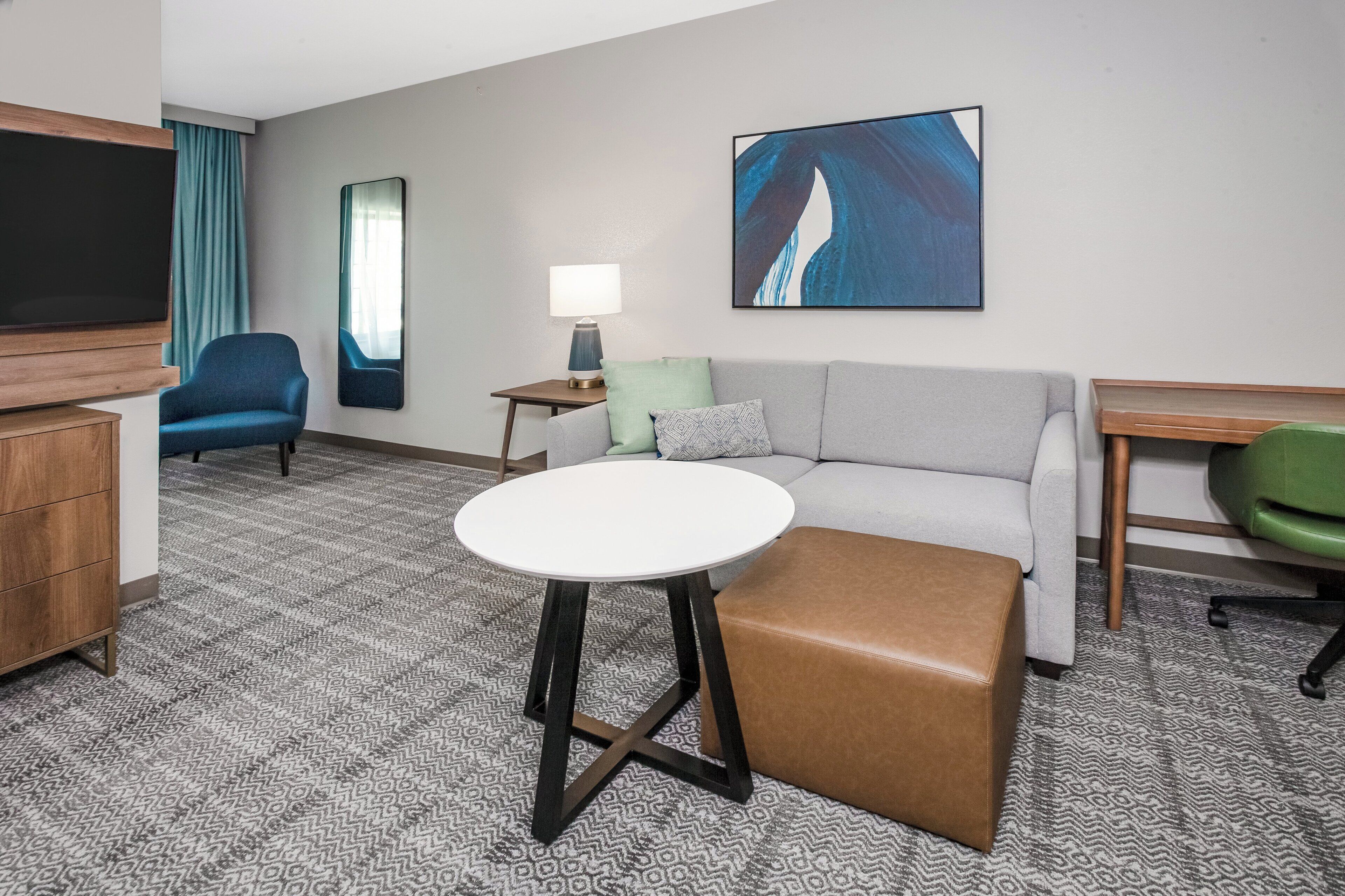 Una habitación de hotel con un sofá gris, un sillón azul, una mesa redonda blanca, un reposapiés marrón, un televisor, una lámpara y un cuadro en la pared.