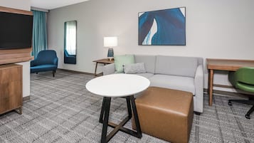 Una habitación de hotel con un sofá gris, un sillón azul, una mesa redonda blanca, un reposapiés marrón, un televisor, una lámpara y un cuadro en la pared.