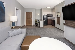 Una habitación de hotel moderna con un sofá gris, un reposapiés marrón, una mesita con una lámpara, un televisor montado en la pared y una zona de cocina con refrigerador, microondas y un taburete.