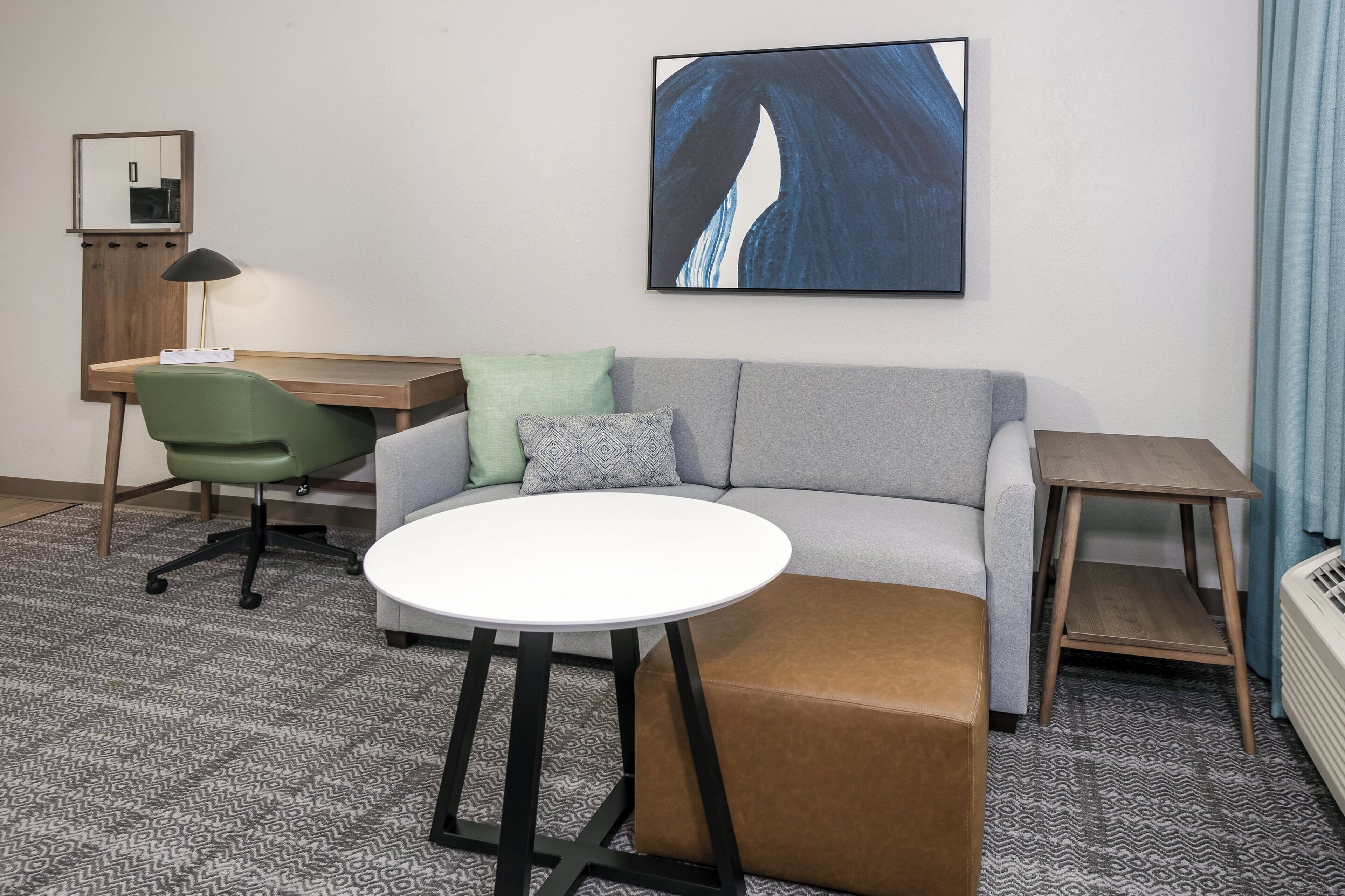 A modern hotel room with a grey sofa, a round white table, a green office chair, a small wooden side table, and a large abstract painting on the wall.