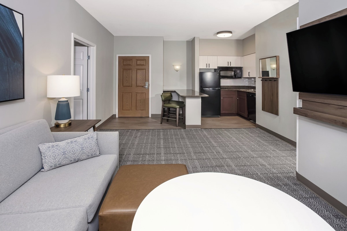 Una habitación de hotel moderna con un sofá gris, un reposapiés marrón, una mesita con una lámpara, un televisor montado en la pared y una zona de cocina con refrigerador, microondas y un taburete.