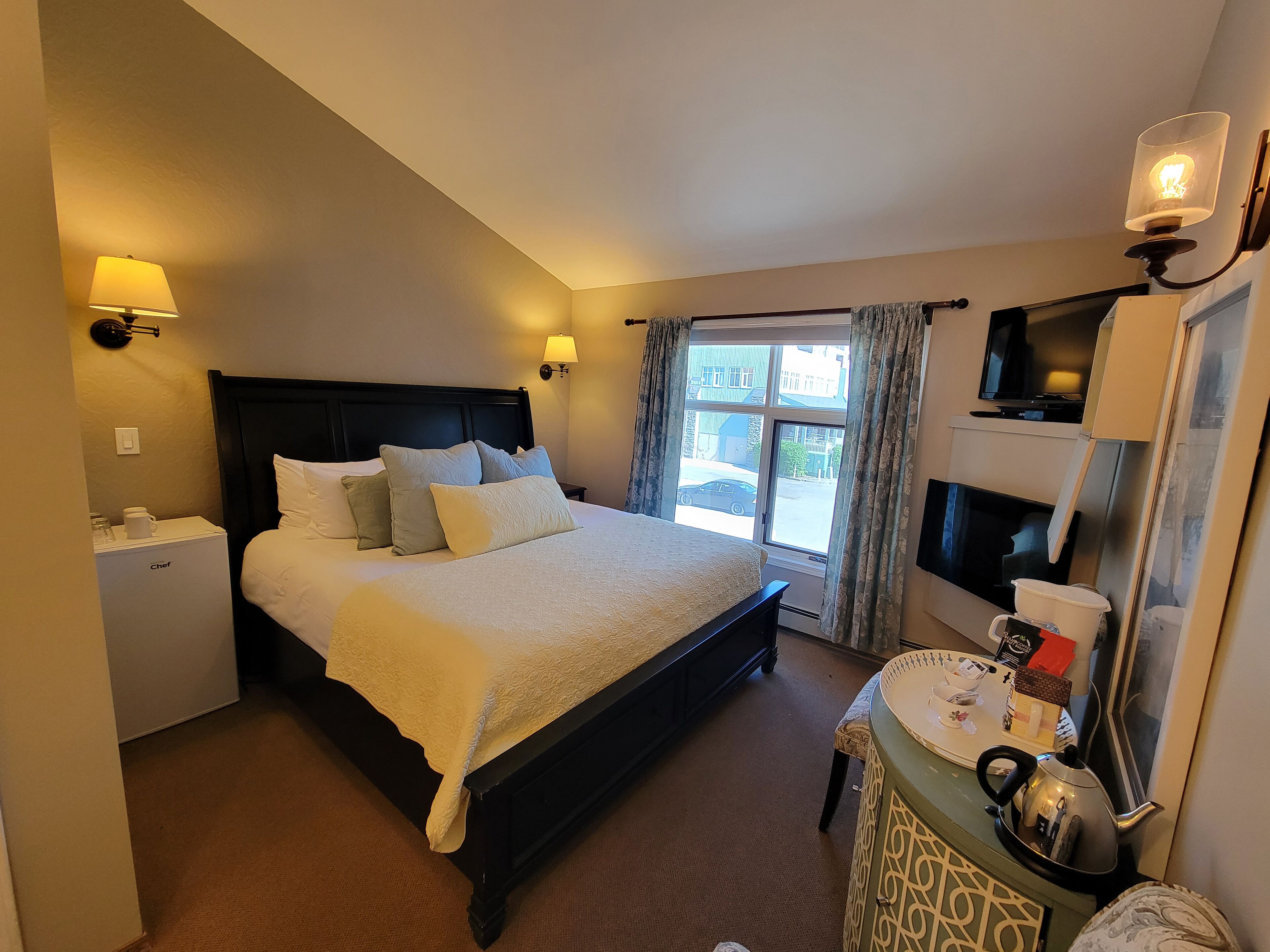 A hotel room with a large bed, bedside lamps, a small refrigerator, a vanity with a kettle, and a television on the wall.