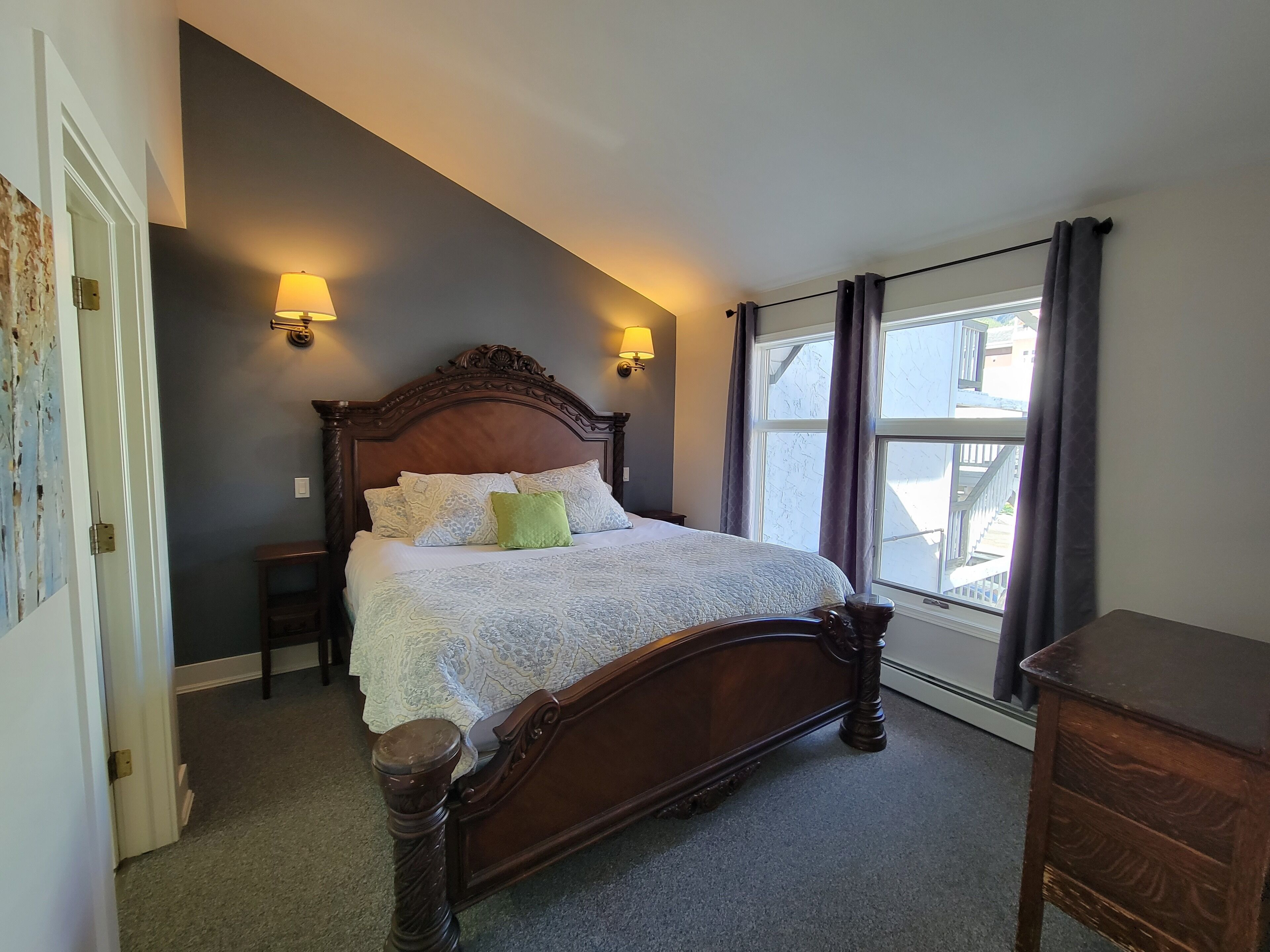 A bedroom with a large bed, two bedside lamps, a window with curtains, and a door with a lock.