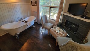 A bathroom with a freestanding bathtub, a chair, a small table with a tray, a television, and a fireplace. - The Georgetown Inn (Canmore)