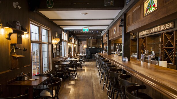 A bar area with wooden flooring, bar stools, and a counter displaying various bottles. - The Georgetown Inn (Canmore)