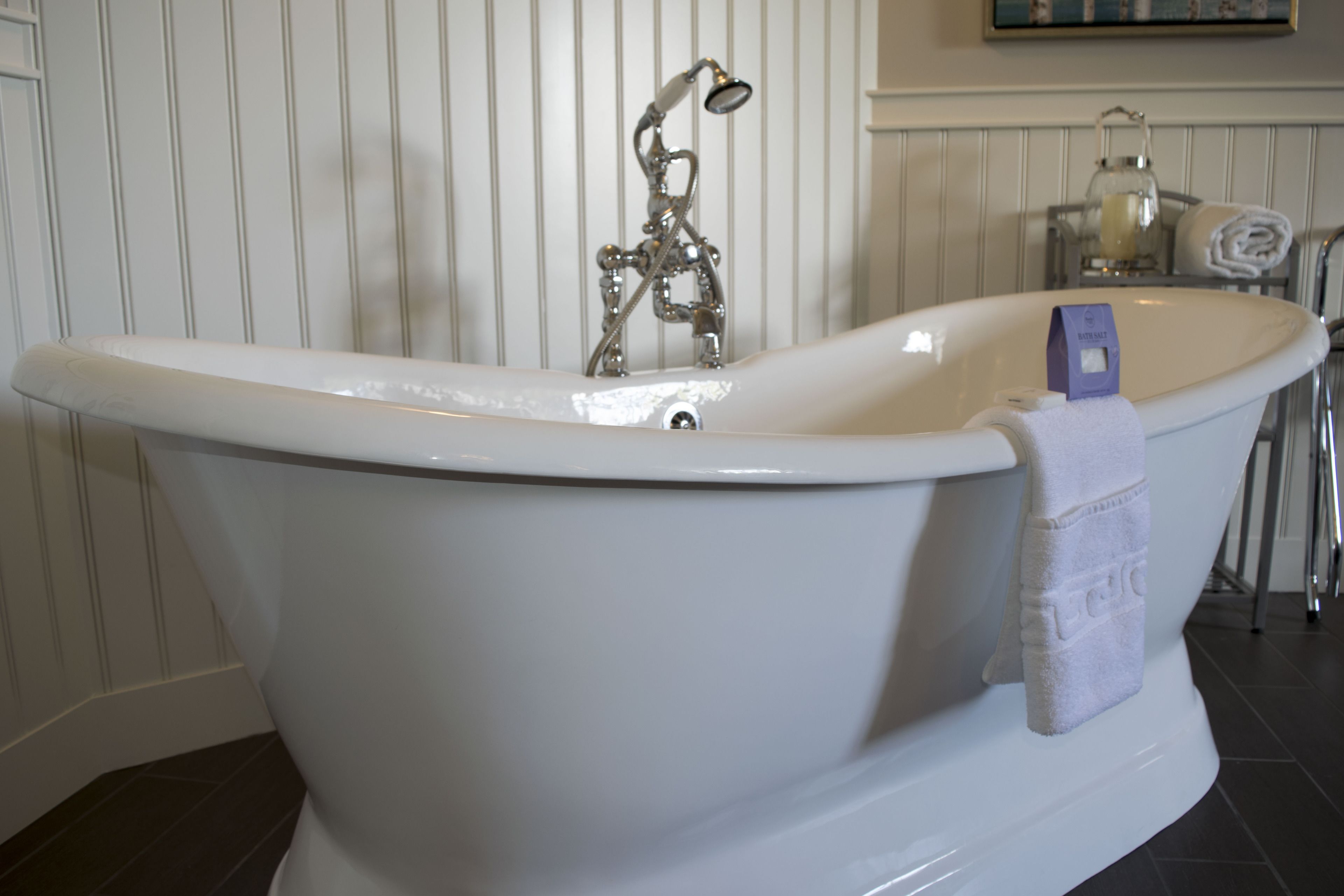 A white clawfoot bathtub with a handheld showerhead, a towel rack, and a glass container on the side.