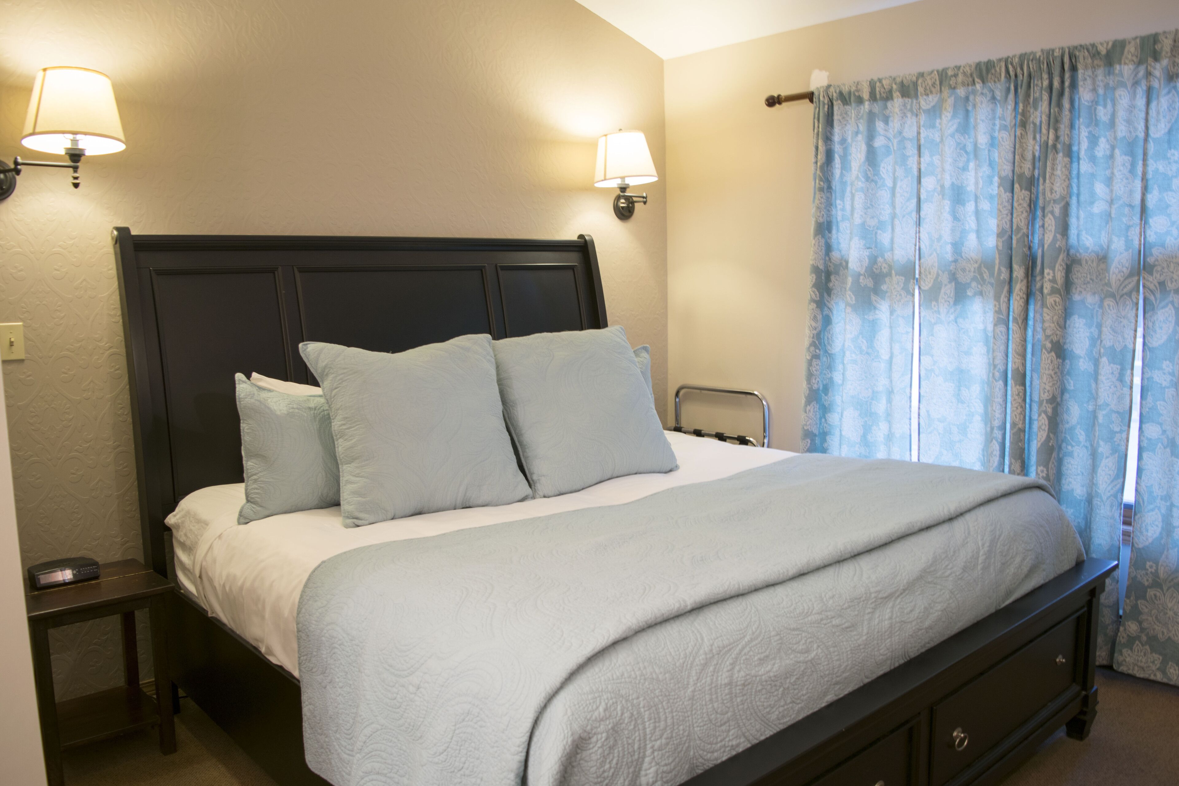 Una cama bien hecha con cabecera oscura, dos lámparas de noche y una ventana con cortinas azules.