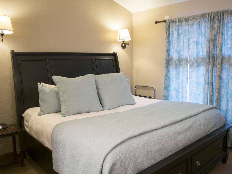 Una cama bien hecha con cabecera oscura, dos lámparas de noche y una ventana con cortinas azules.