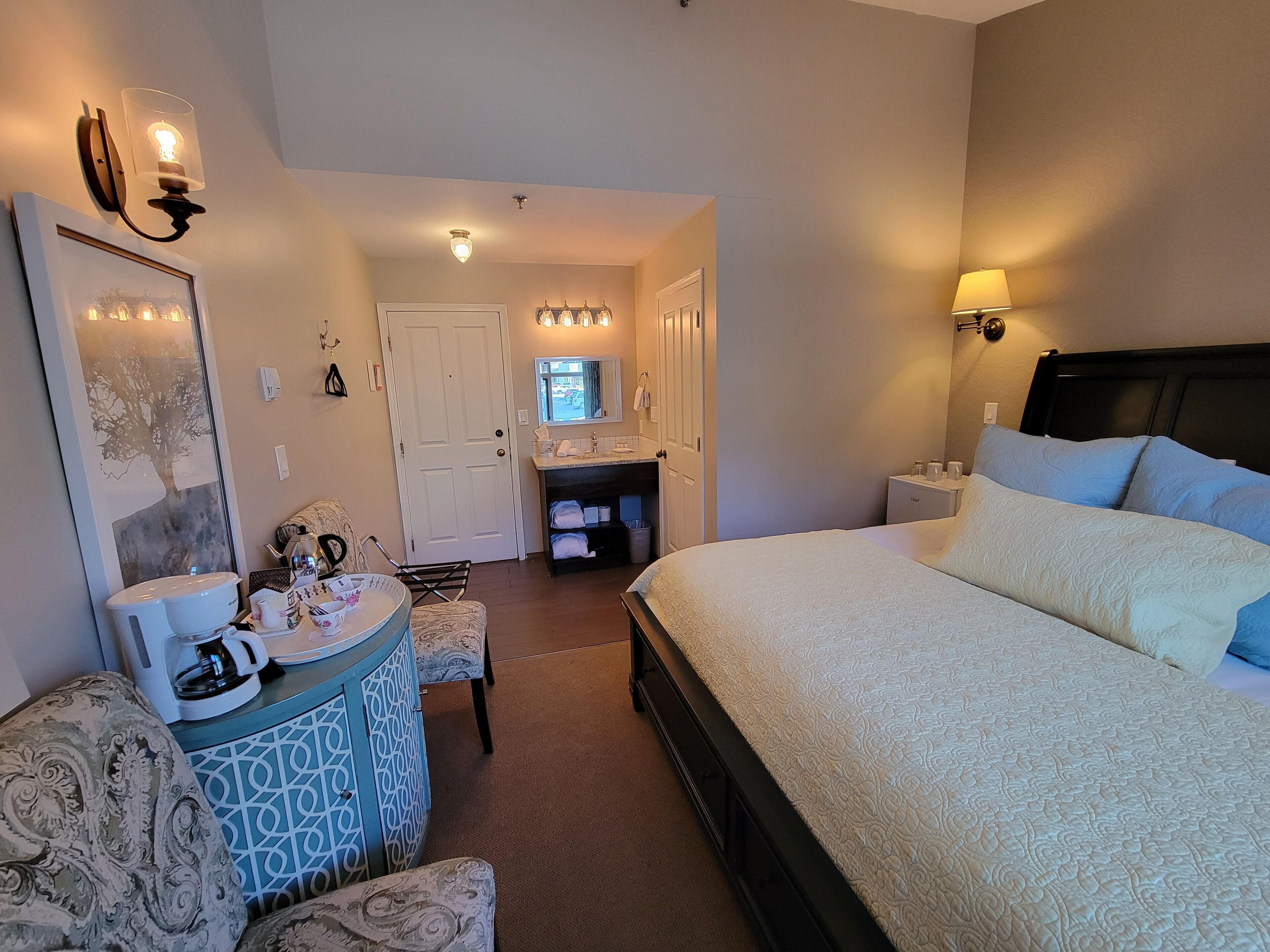 Habitación de hotel con cama, escritorio con cafetera, una silla, baño con espejo y lavamanos, y puerta que da a otra habitación.