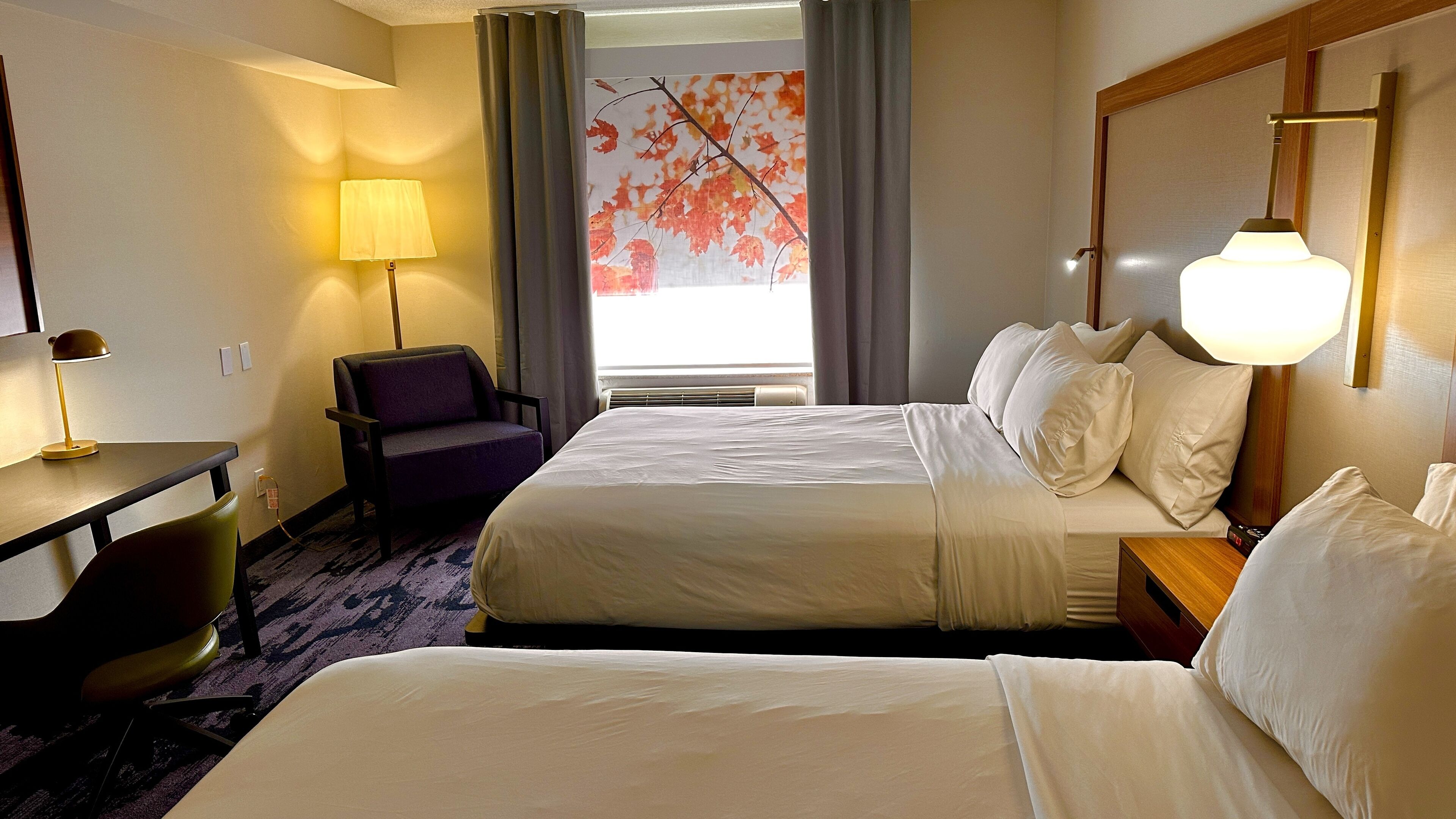 A hotel room with two beds, a desk with a lamp, a chair, and a painting of autumn leaves on the wall.