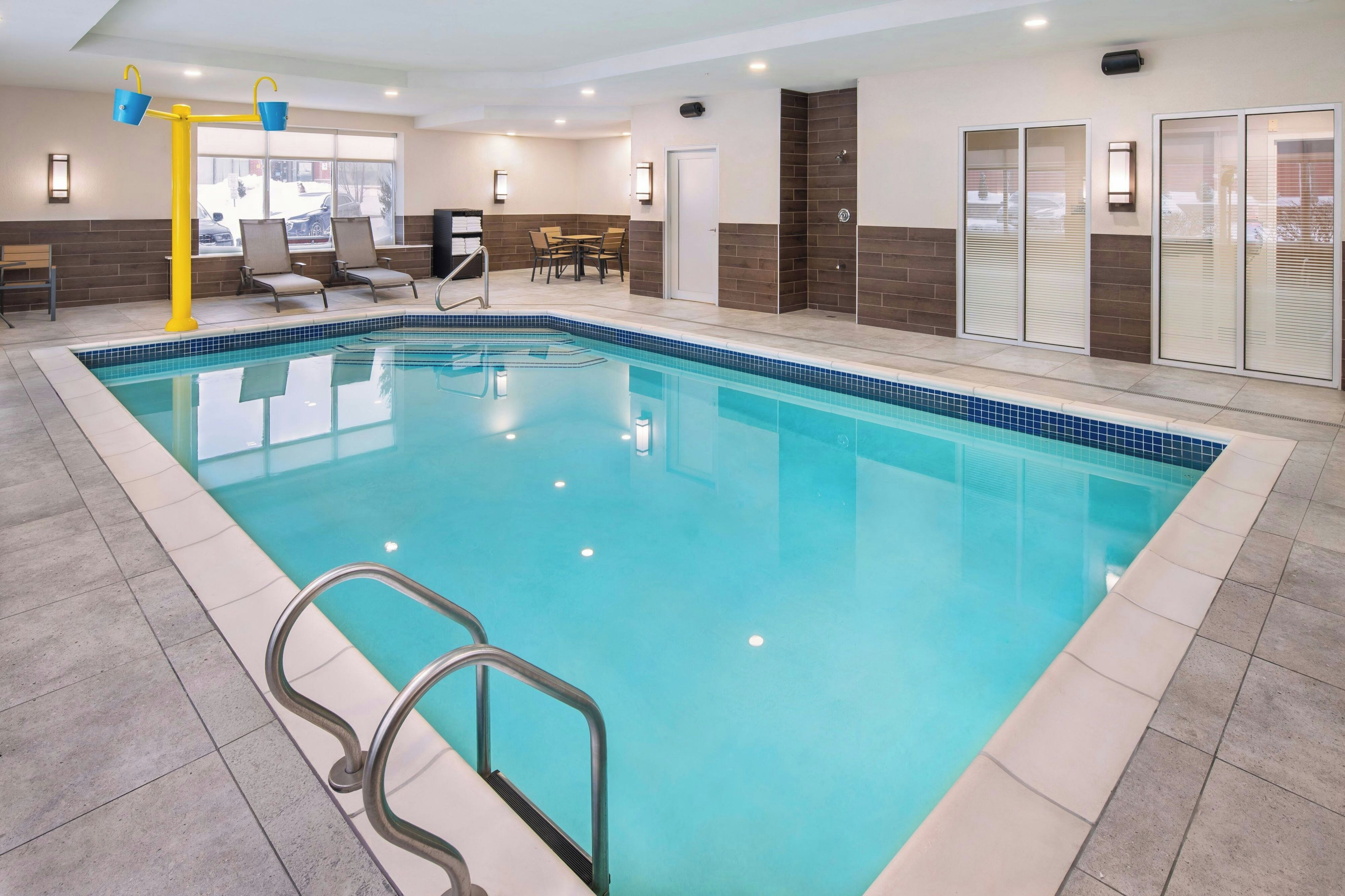 Una piscina con borde de azulejos, un puesto de socorrista amarillo y una zona de estar con sillas.