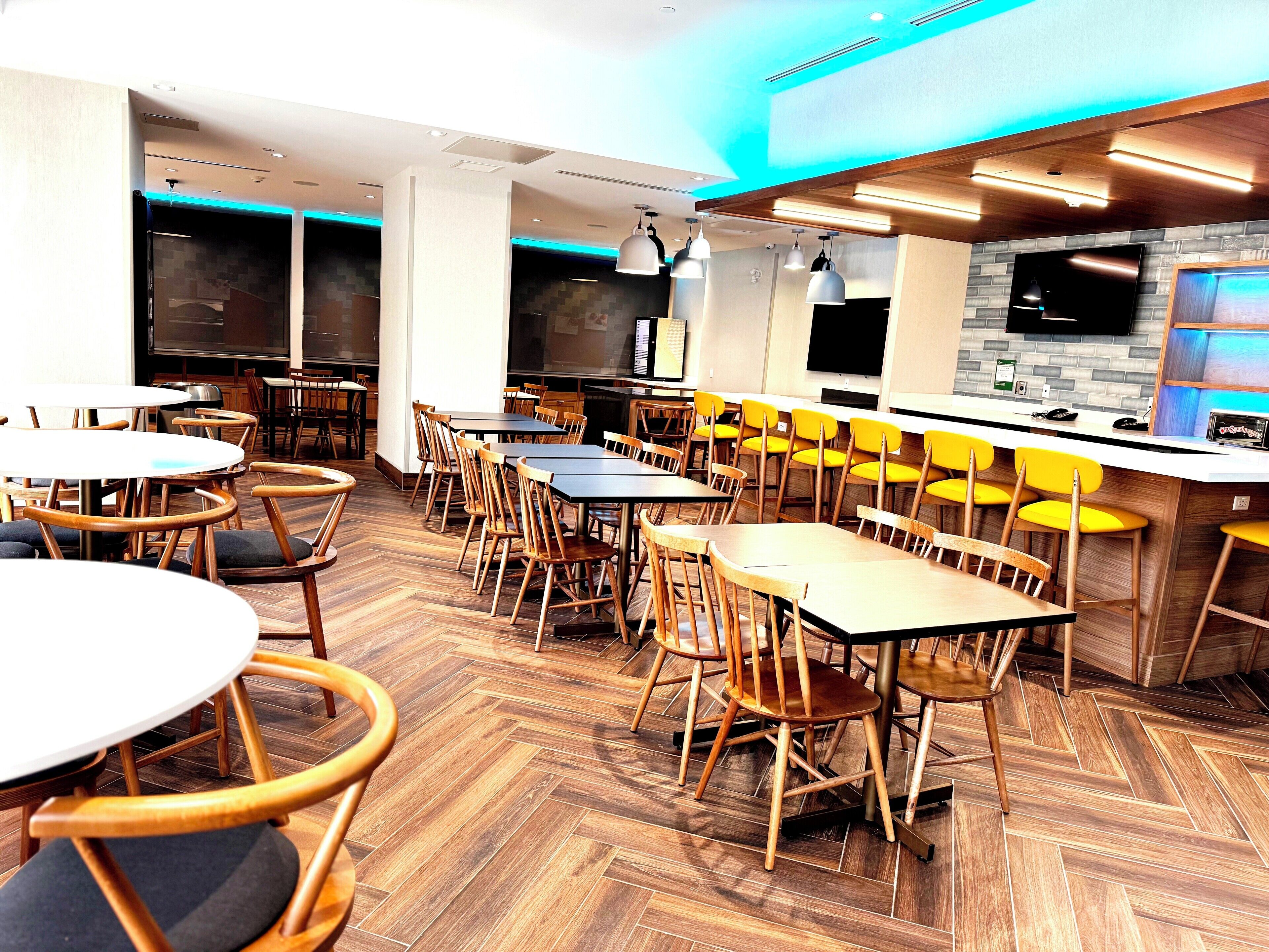 A modern restaurant with wooden tables and chairs, a bar area, and a large TV screen.