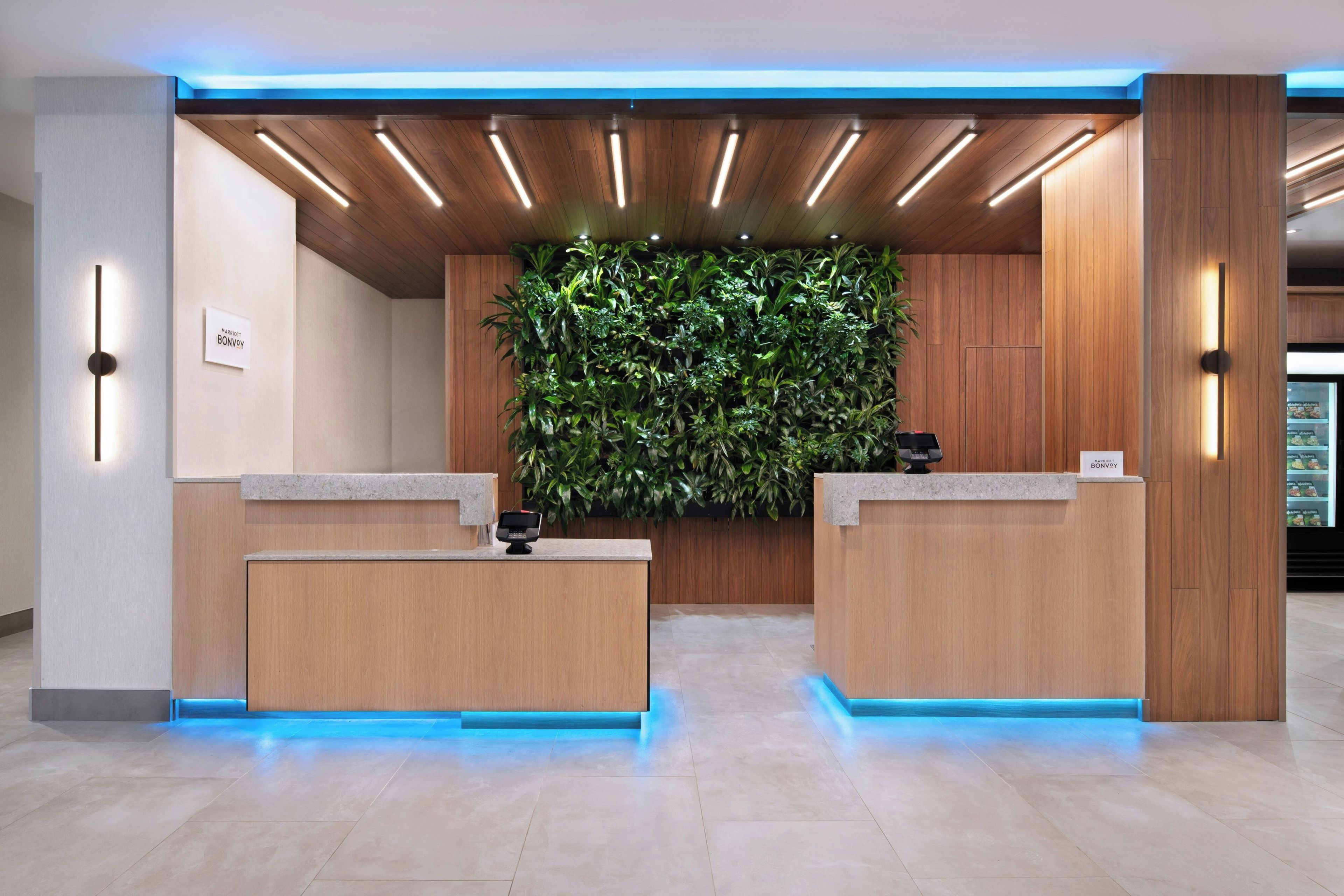 Een hotel receptie met twee houten balies, een groene verticale tuin en een blauw verlicht plafond.