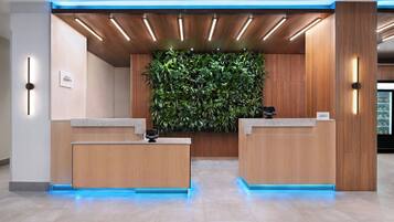 Een hotel receptie met twee houten balies, een groene verticale tuin en een blauw verlicht plafond.