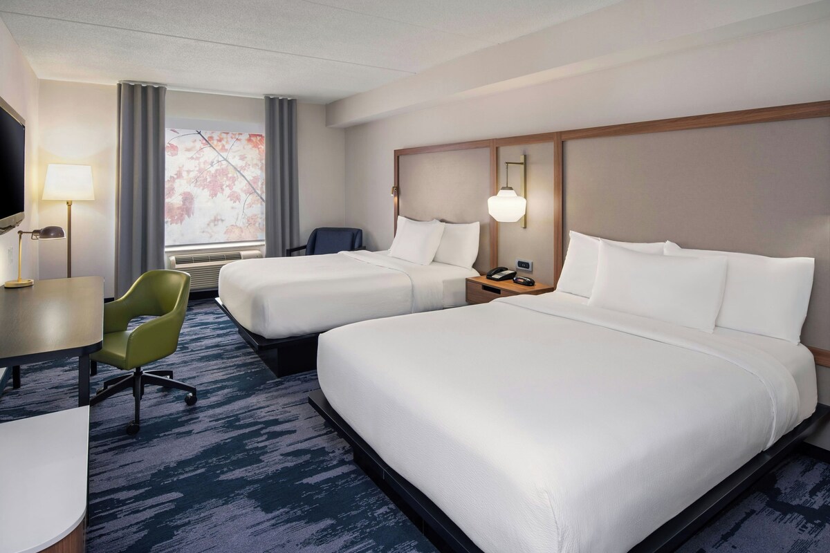 Une chambre d’hôtel avec deux lits, un bureau, une chaise, une télévision et une fenêtre donnant sur des arbres.