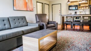 Una sala de estar moderna con un sofá gris, un sillón de cuero negro y una mesa de centro de madera. Se ve un área de cocina con microondas, estufa y asientos al fondo.