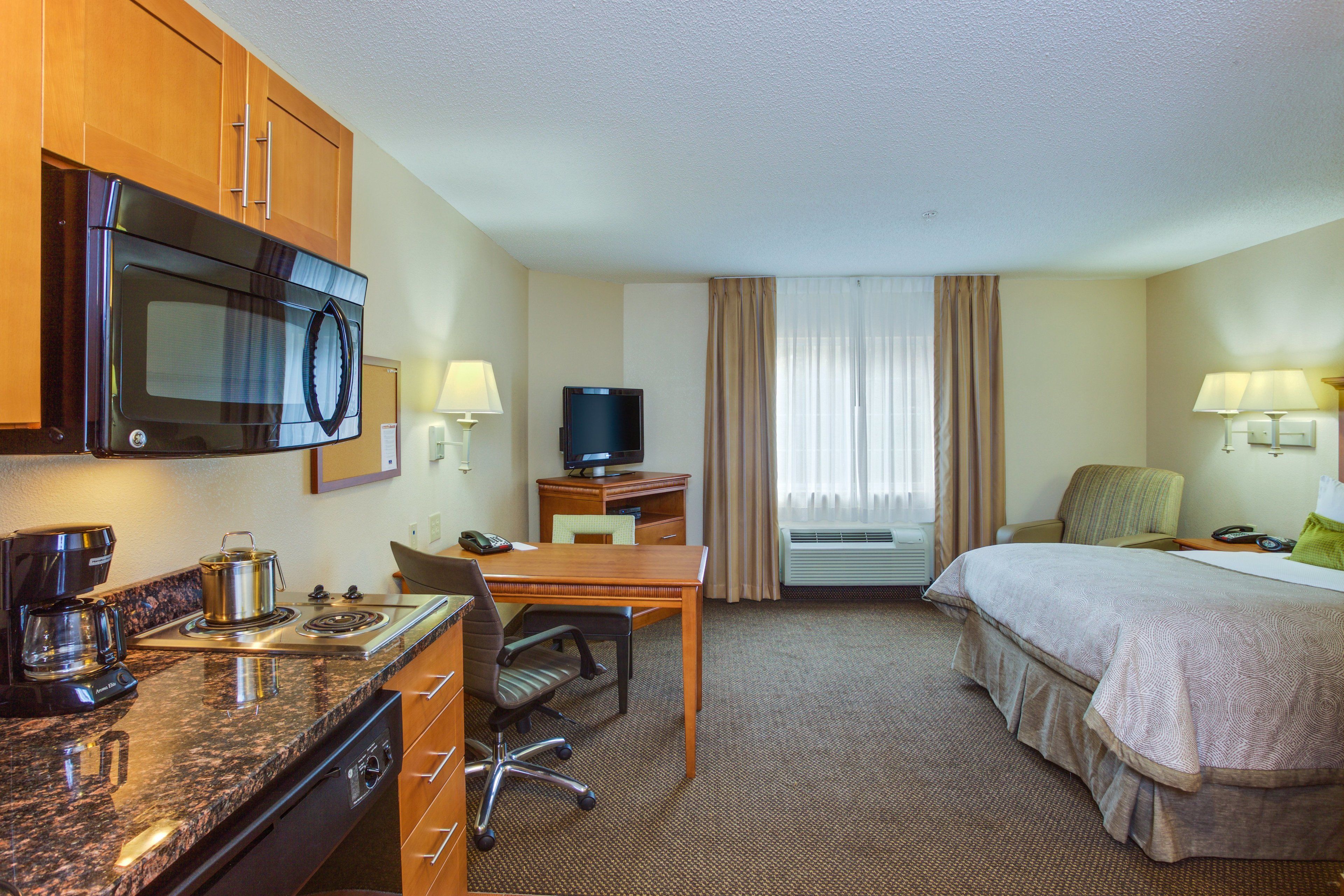 A hotel room with a kitchenette, a bed, a desk with a chair, a television, and a window with curtains.