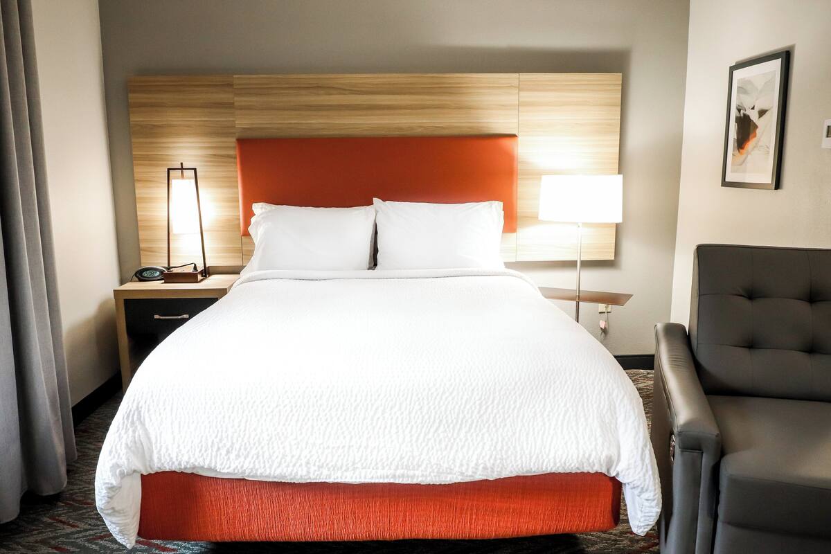 A hotel room with a large bed, a nightstand with a lamp, a chair, and a framed picture on the wall.