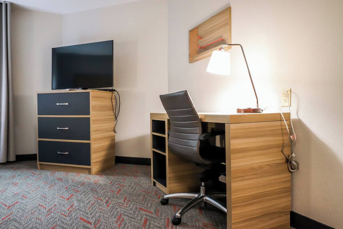 A hotel room with a wooden desk, a black office chair, a television on top of a dresser, and a lamp on the desk.