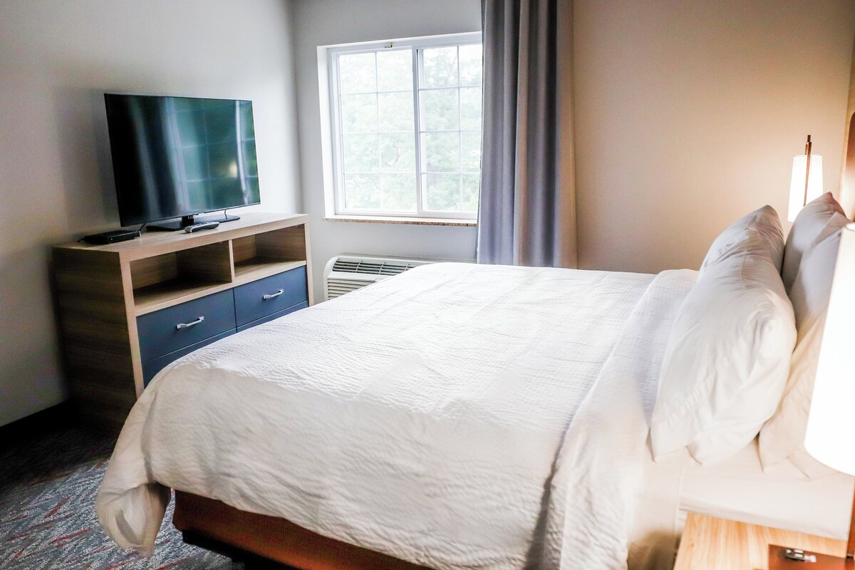 Habitación de hotel con cama, televisión sobre un mueble bajo, ventana con cortinas y mesita de noche con lámpara.