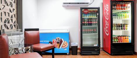 Uma área de estar com mesa, cadeiras, máquina de vendas e um refrigerador.