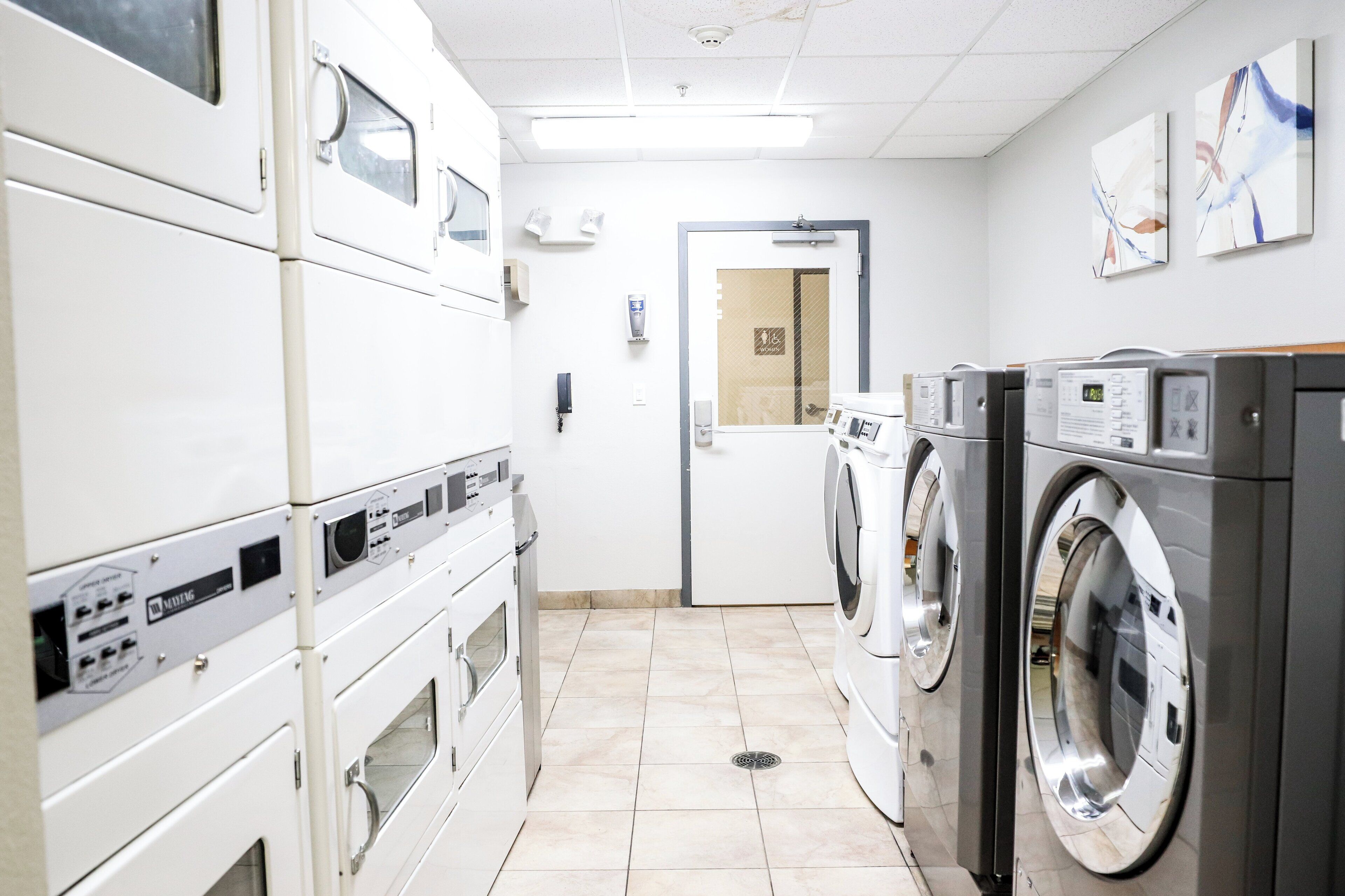 Une buanderie équipée de plusieurs machines à laver et de plusieurs sèche-linge, avec un sol carrelé et une porte donnant sur une autre pièce.
