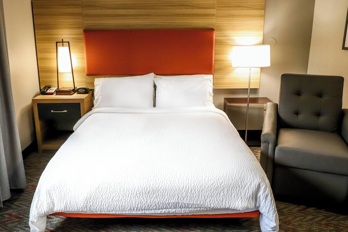 A hotel room with a large bed, a nightstand with a phone and lamp, a chair, and a small table.