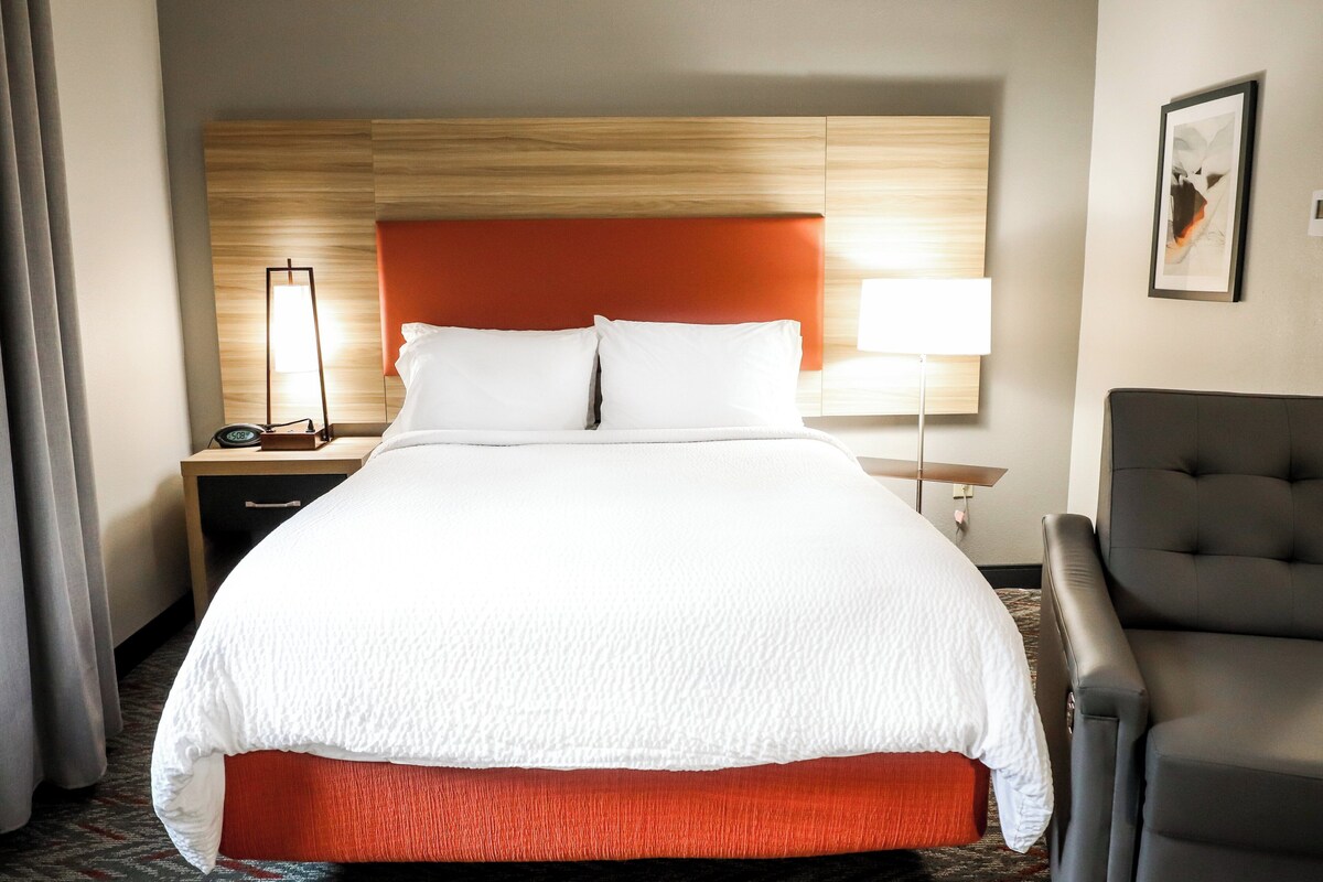 Un cuarto de hotel con una cama grande, una mesita de noche con lámpara, una silla y un cuadro en la pared.