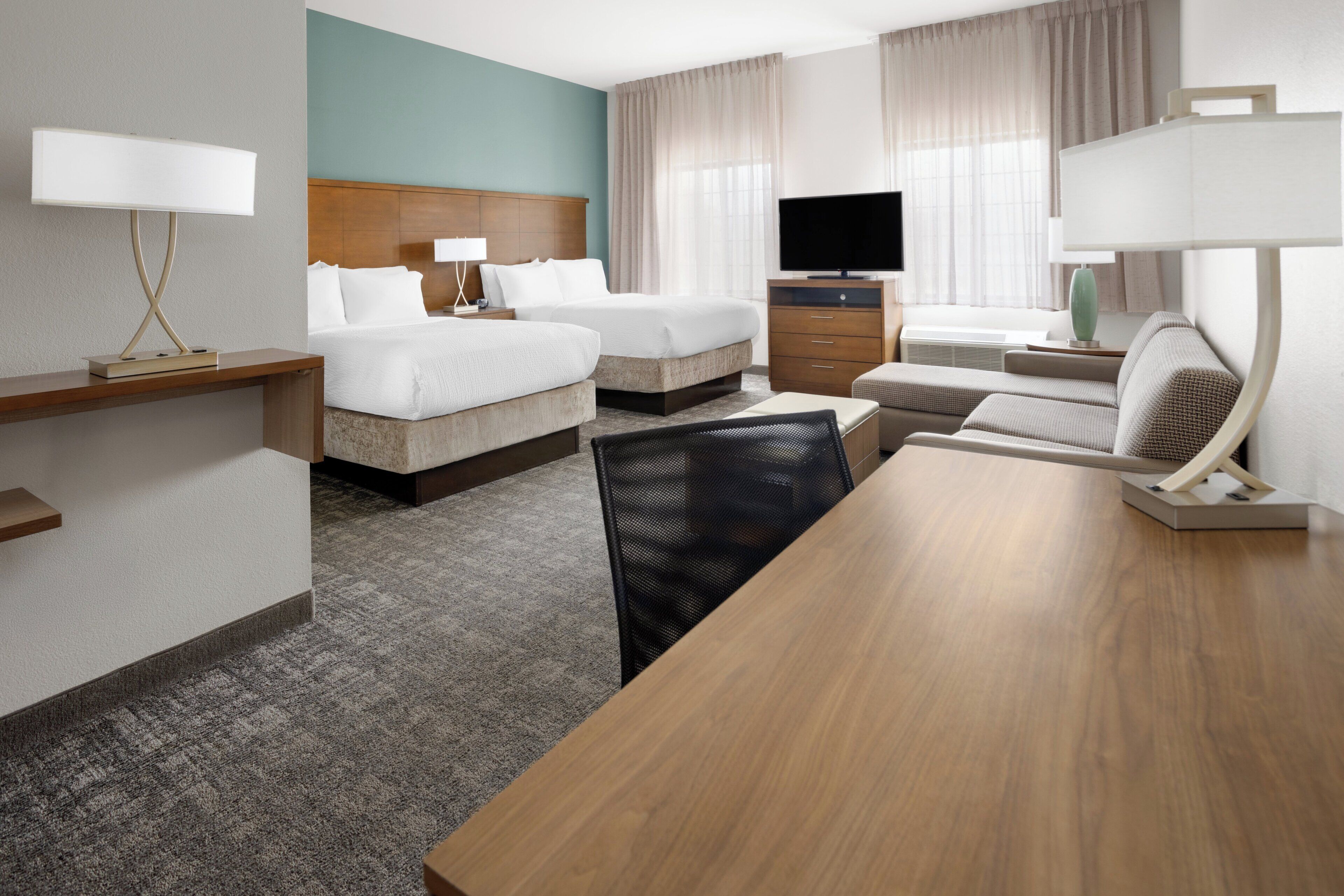 A hotel room with two beds, a TV, a desk with a lamp, a chair, and a large wooden table.