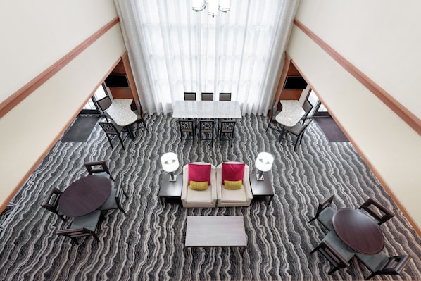A hotel lobby with a central seating area featuring a sofa set, two side tables, and a coffee table. There are four chairs around the sofa, and a dining table with chairs on the left. The floor has a patterned carpet, and the walls are plain with a wooden trim.