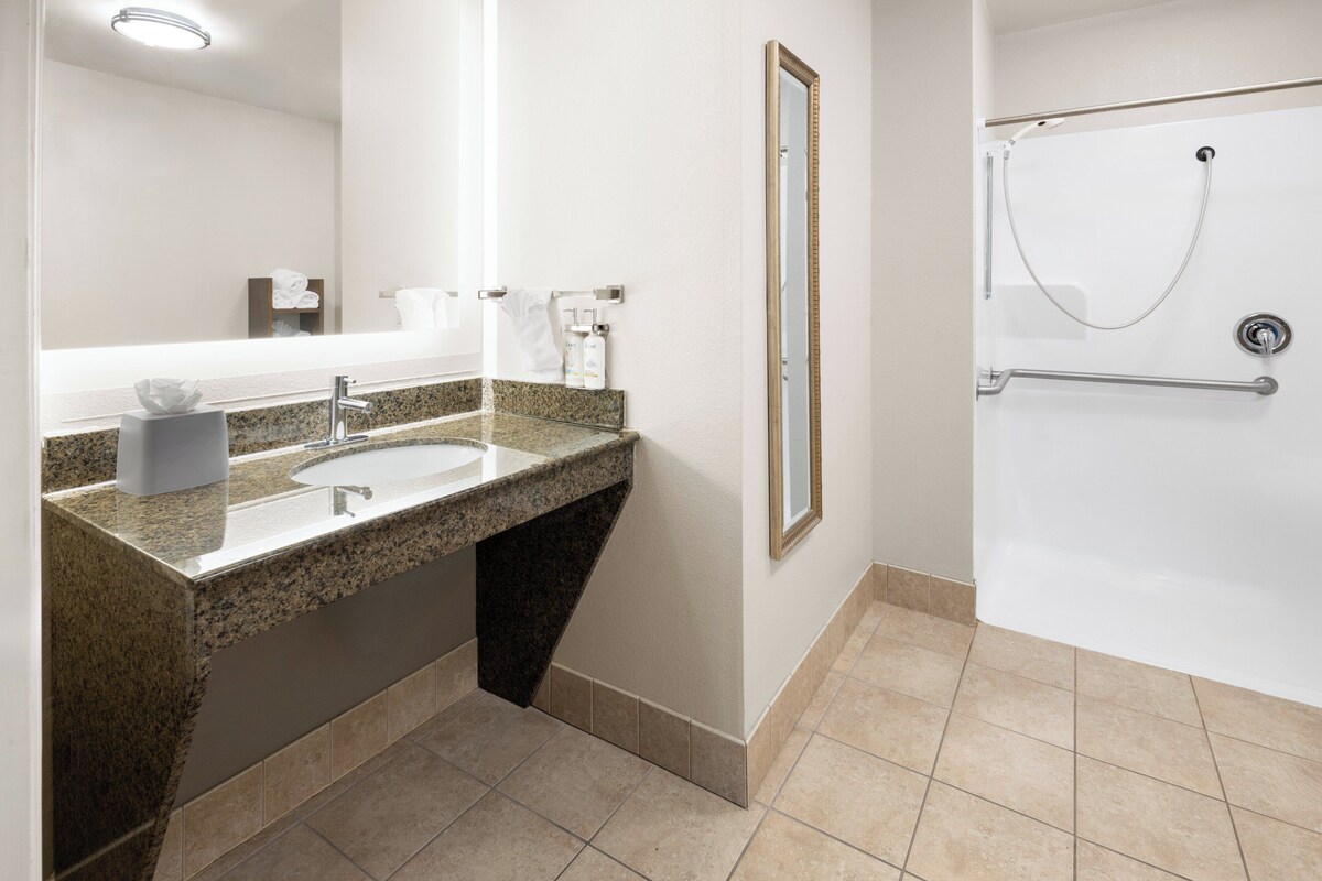 A modern bathroom with a granite countertop sink, a large mirror, and a walk-in shower with a grab bar.