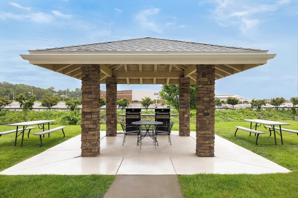 A gazebo with a stone base and a tiled roof, surrounded by picnic tables and a barbecue grill.
