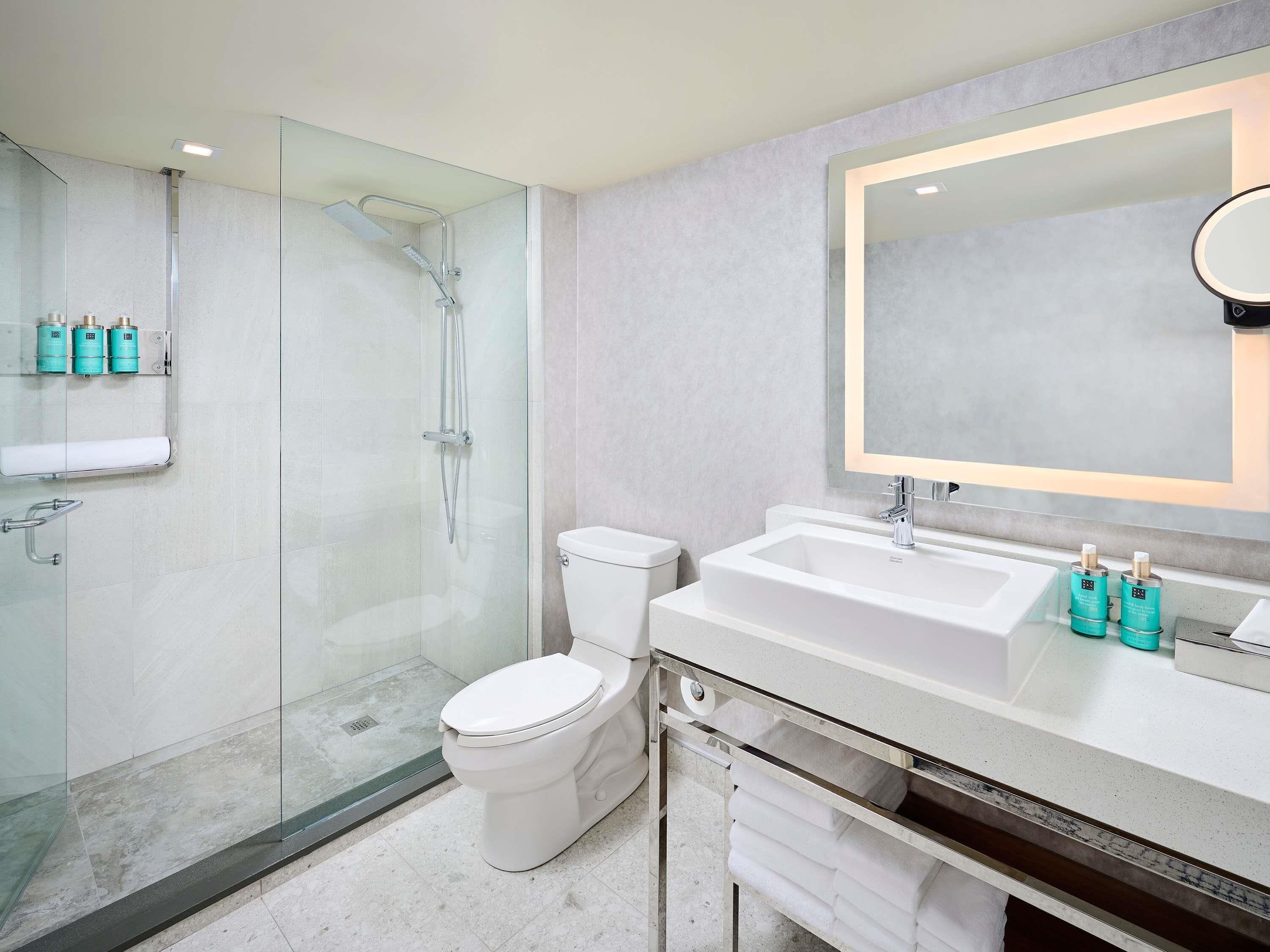 A modern bathroom with a glass-enclosed shower, a white toilet, a large mirror, and a countertop with toiletries.