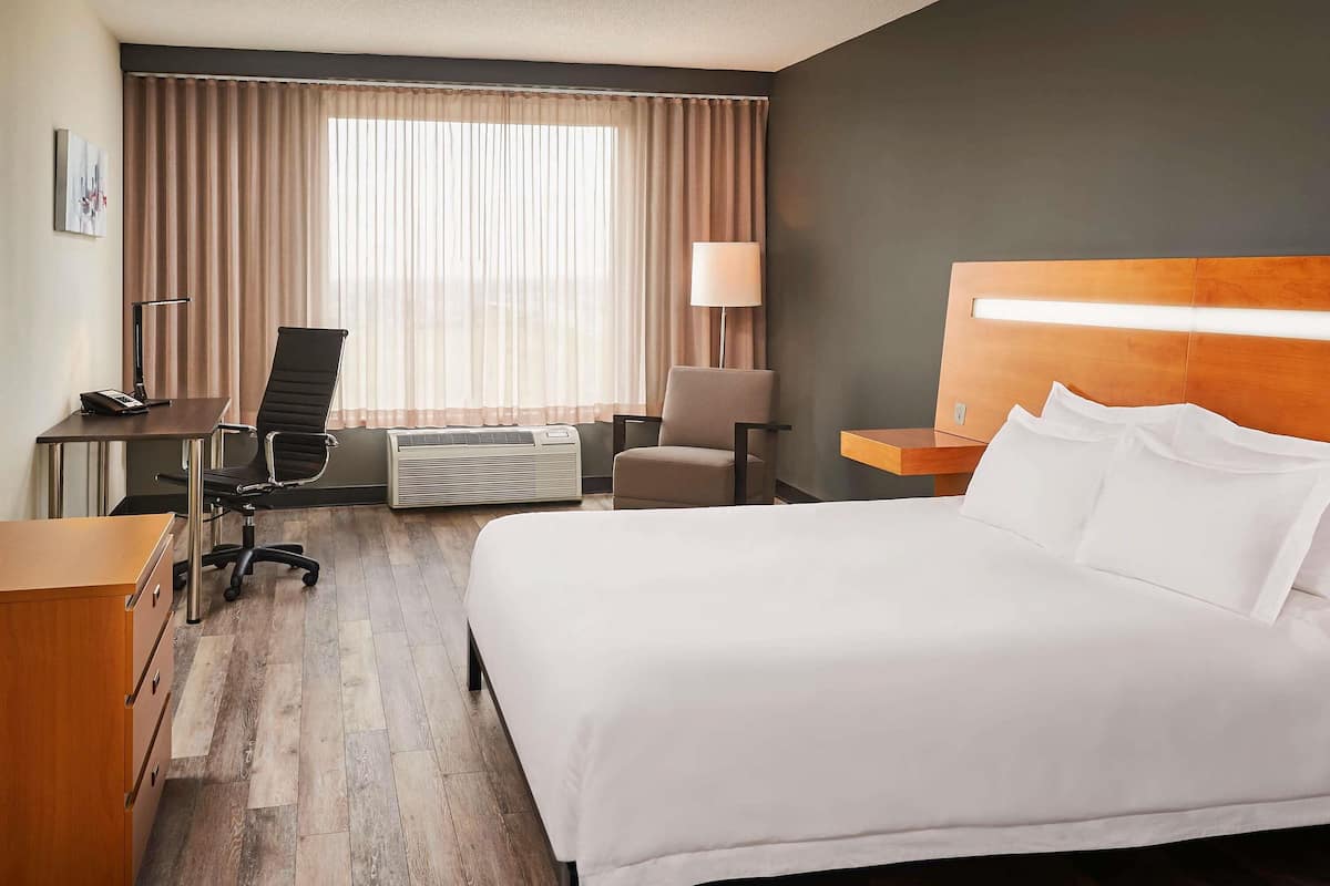 Habitación de hotel con cama, escritorio, silla, equipo de aire acondicionado, lámpara y vistas al exterior a través de cortinas.