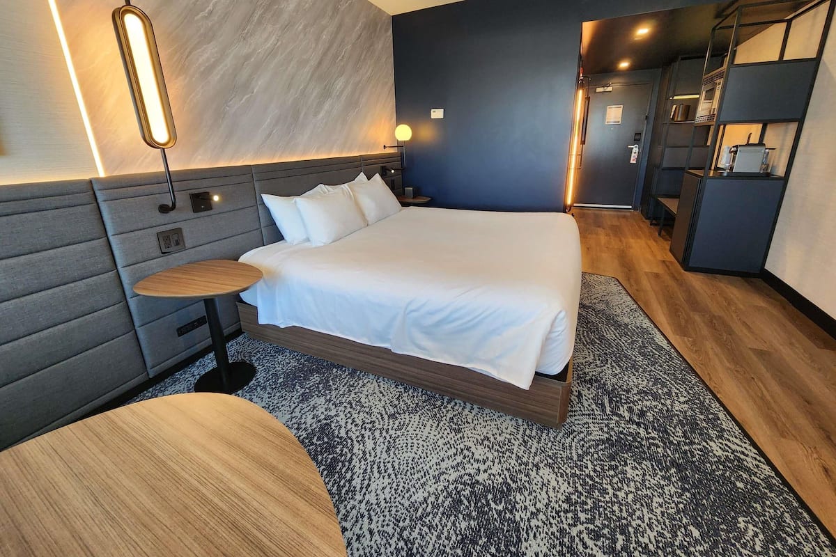 Una habitación de hotel moderna con una cama grande, una mesita de noche, una lámpara de pared y una pequeña zona de escritorio.