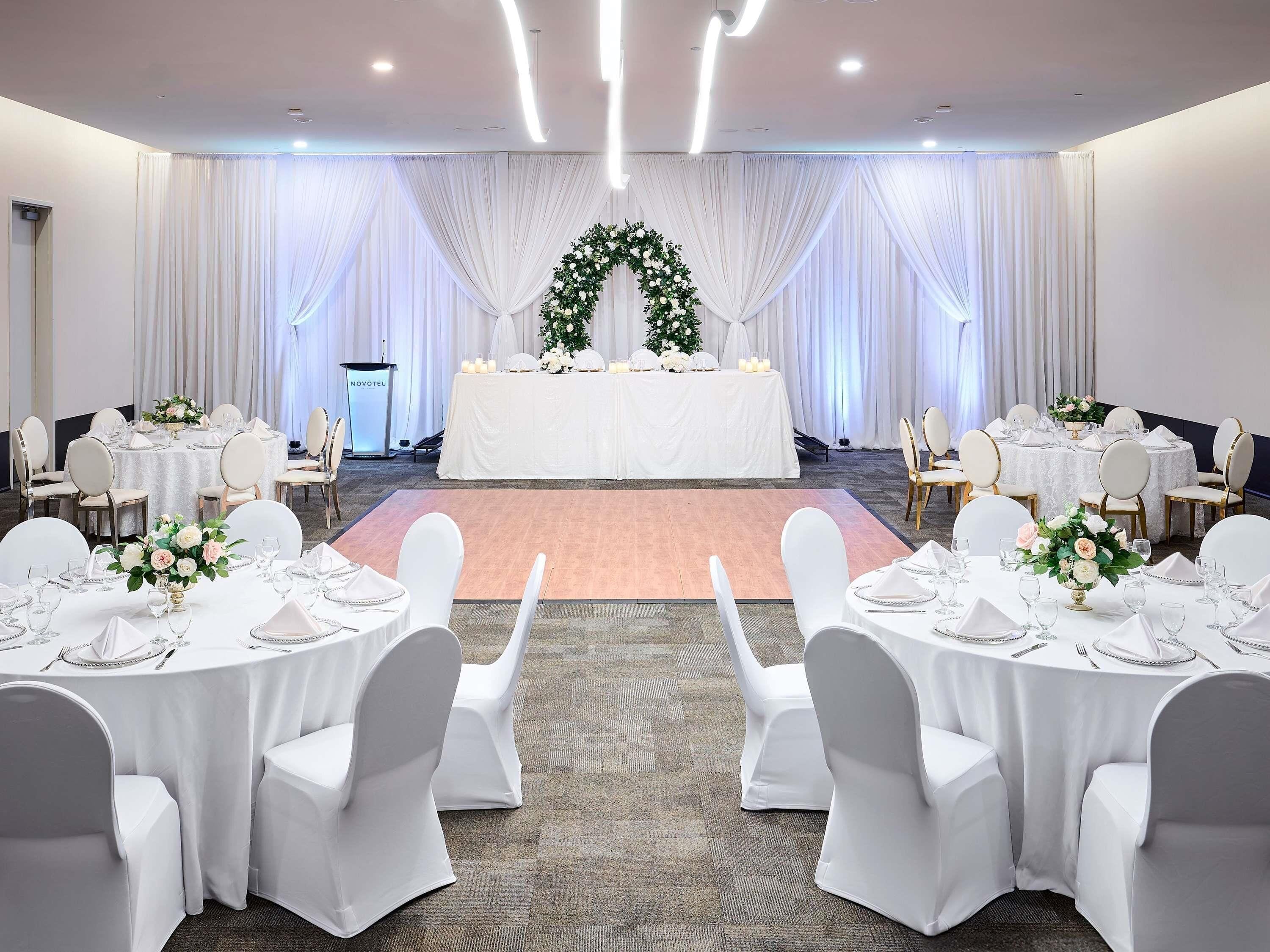 A banquet hall with round tables, white chairs, a stage with a floral backdrop, and a podium.
