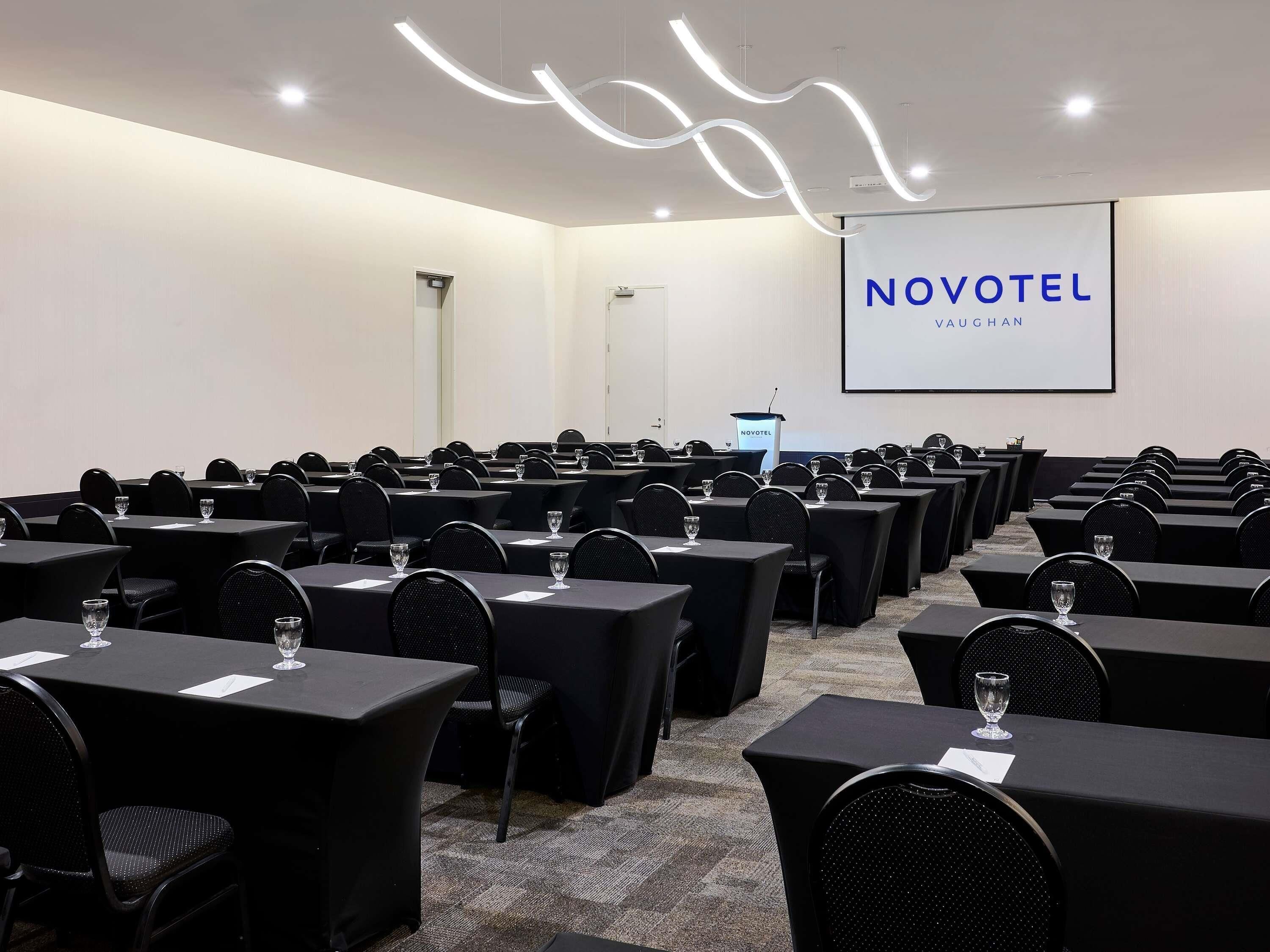 A conference room with rows of tables and chairs, a podium, and a screen displaying 'NOVOTEL VAUGAN'.