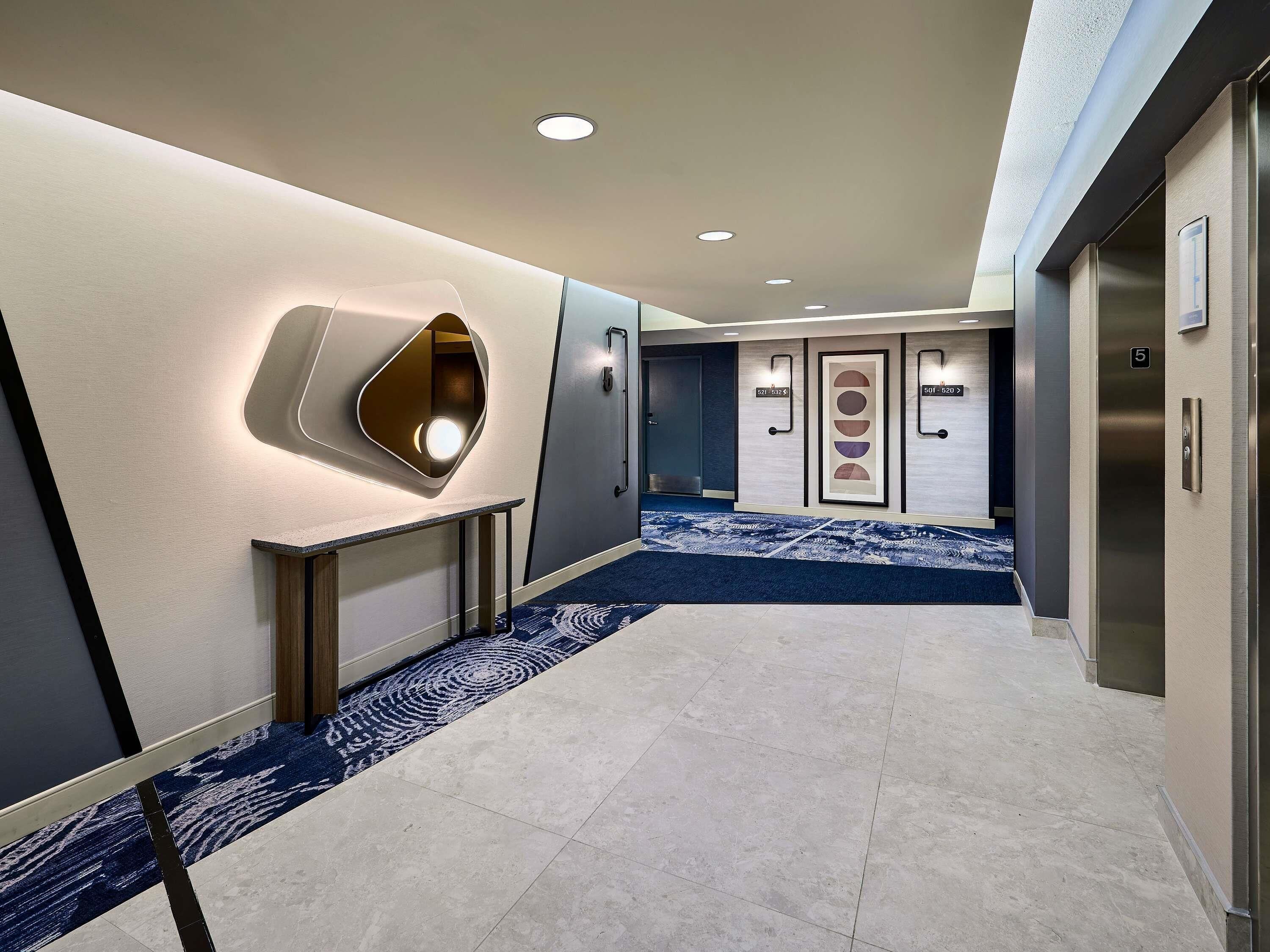 Un pasillo moderno de hotel con alfombra azul, iluminación empotrada y un panel informativo en la pared.