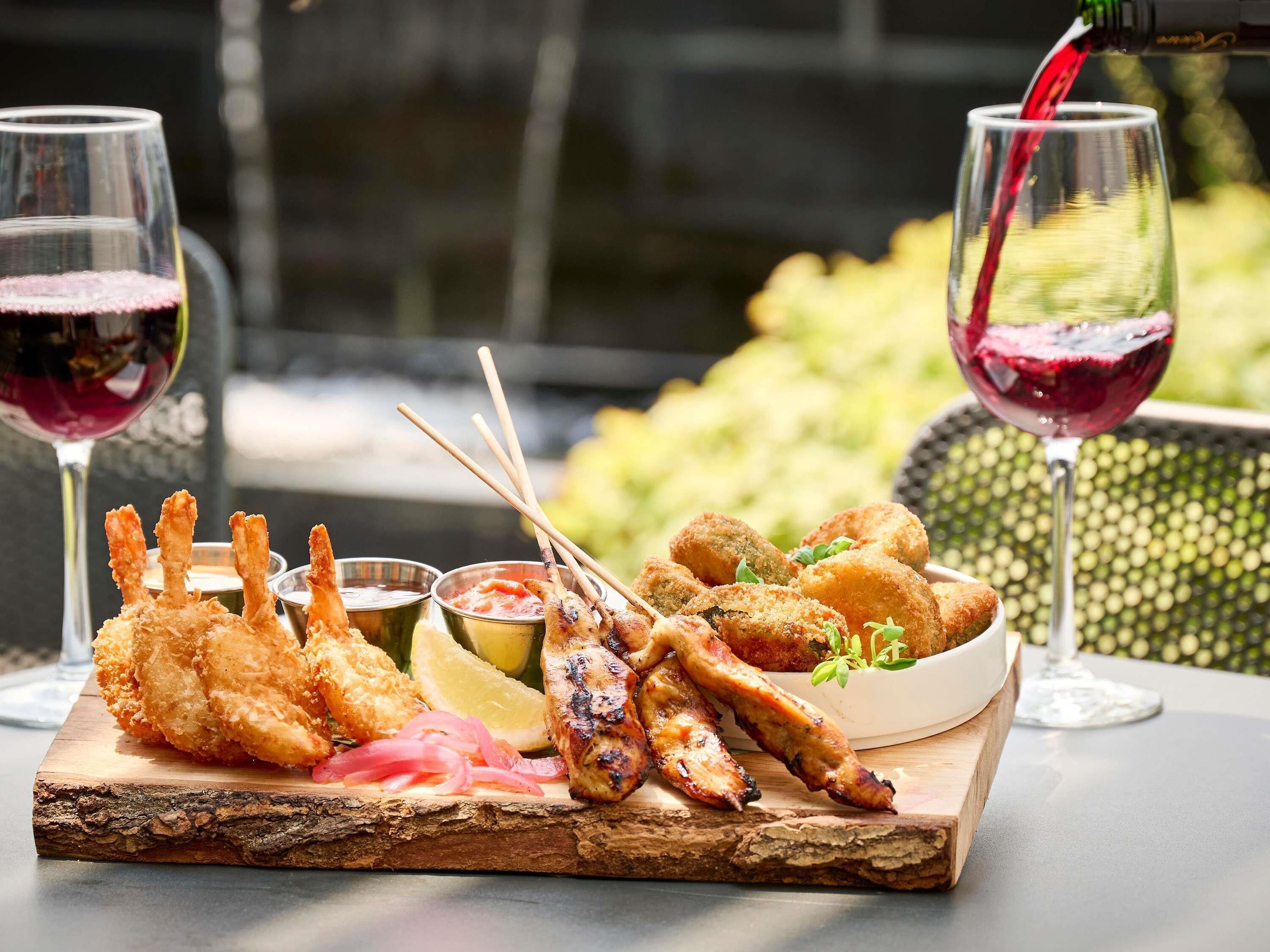 Un tagliere di legno con pesce fritto, fettine di limone e salse. Sul tavolo ci sono due bicchieri di vino rosso.
