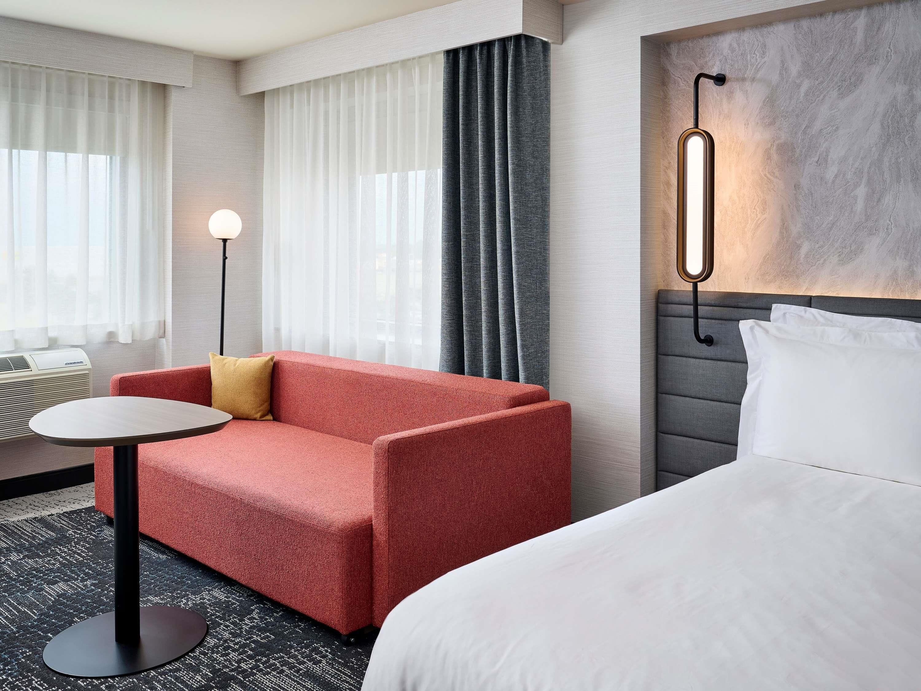Une chambre d’hôtel avec un canapé rouge, une table ronde, un lit et un luminaire fixé au mur.
