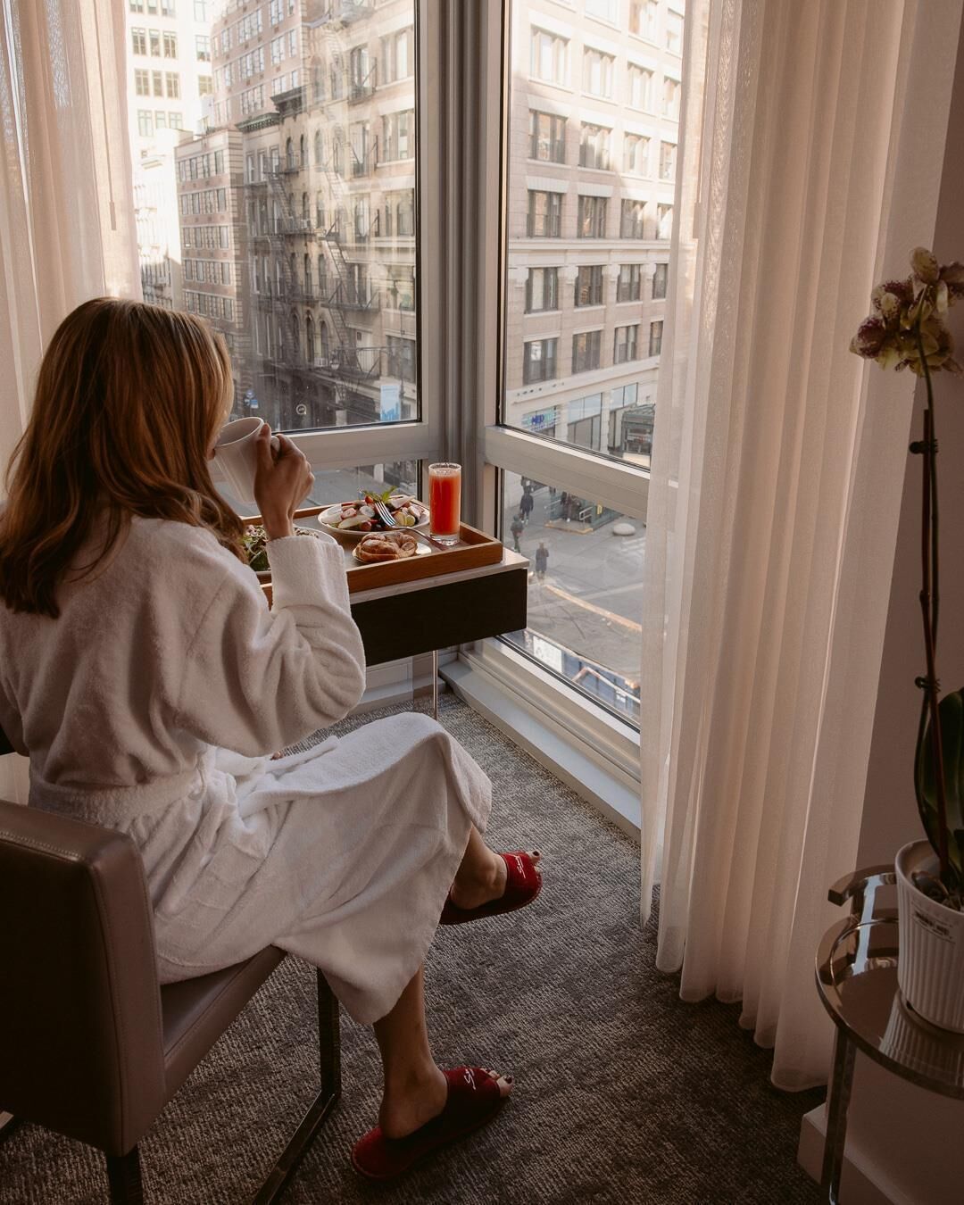 A person in a robe sitting by a window with a tray of food and drink.