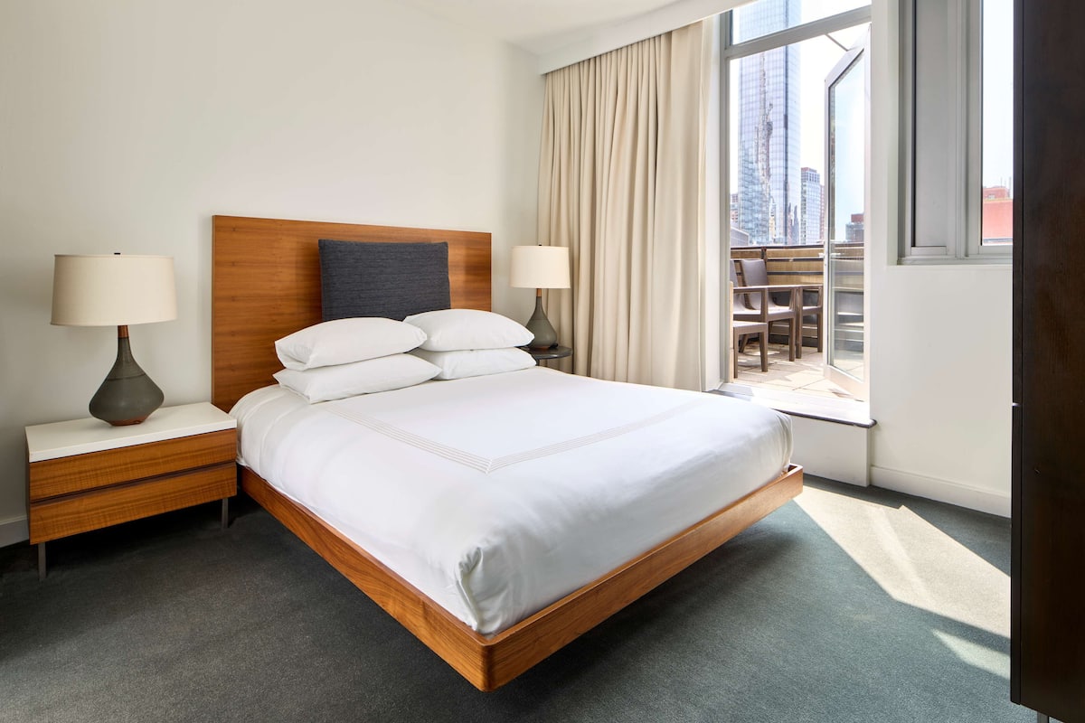 A hotel room with a wooden headboard, a bed with white linens, two bedside lamps, a nightstand, and a view of the city through large windows.