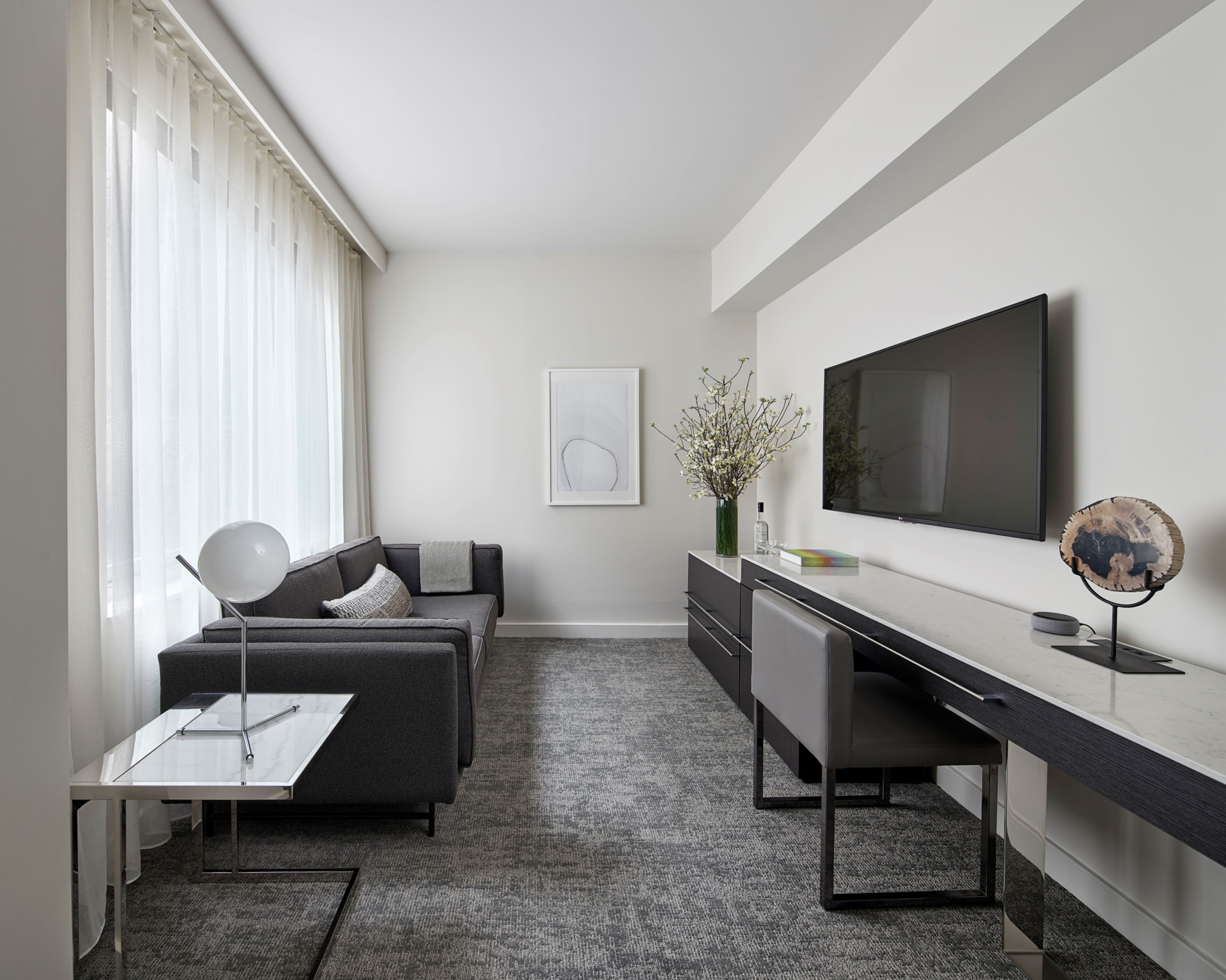 Une chambre d’hôtel moderne avec un canapé gris, un bureau, un téléviseur à écran plat et une grande fenêtre agrémentée de rideaux légers.
