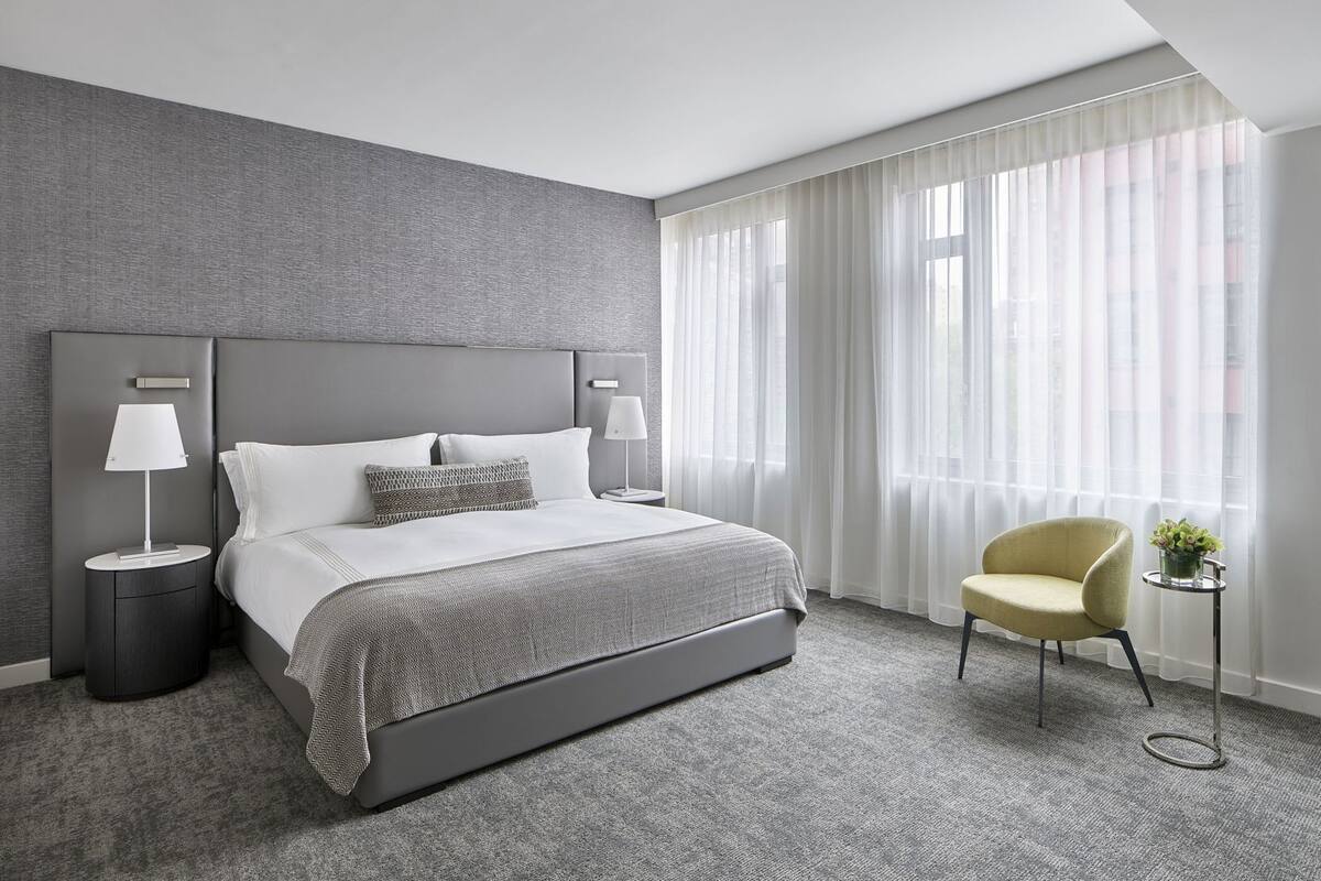 A modern hotel room with a large bed, two bedside tables with lamps, a small armchair, and a view of the city through sheer curtains.