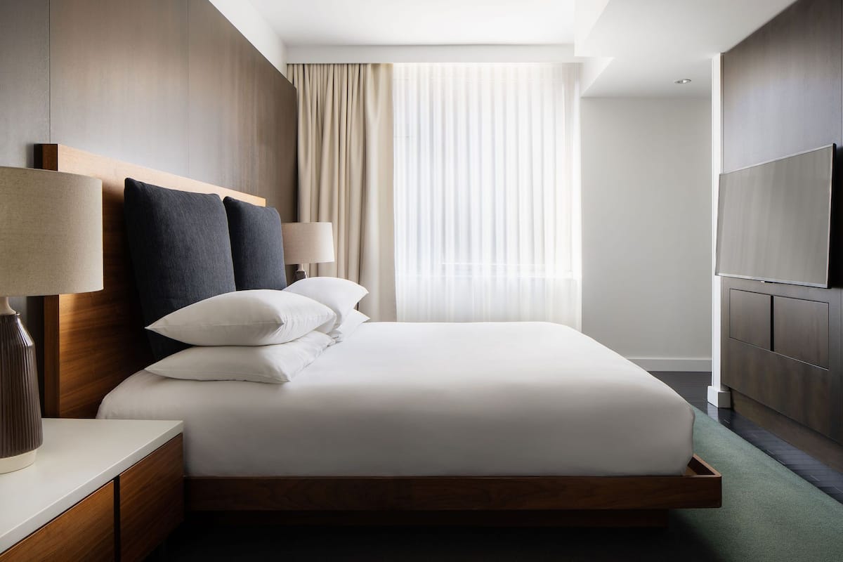 A modern hotel room with a large bed, wooden headboard, a flat-screen TV, and a large window with curtains.