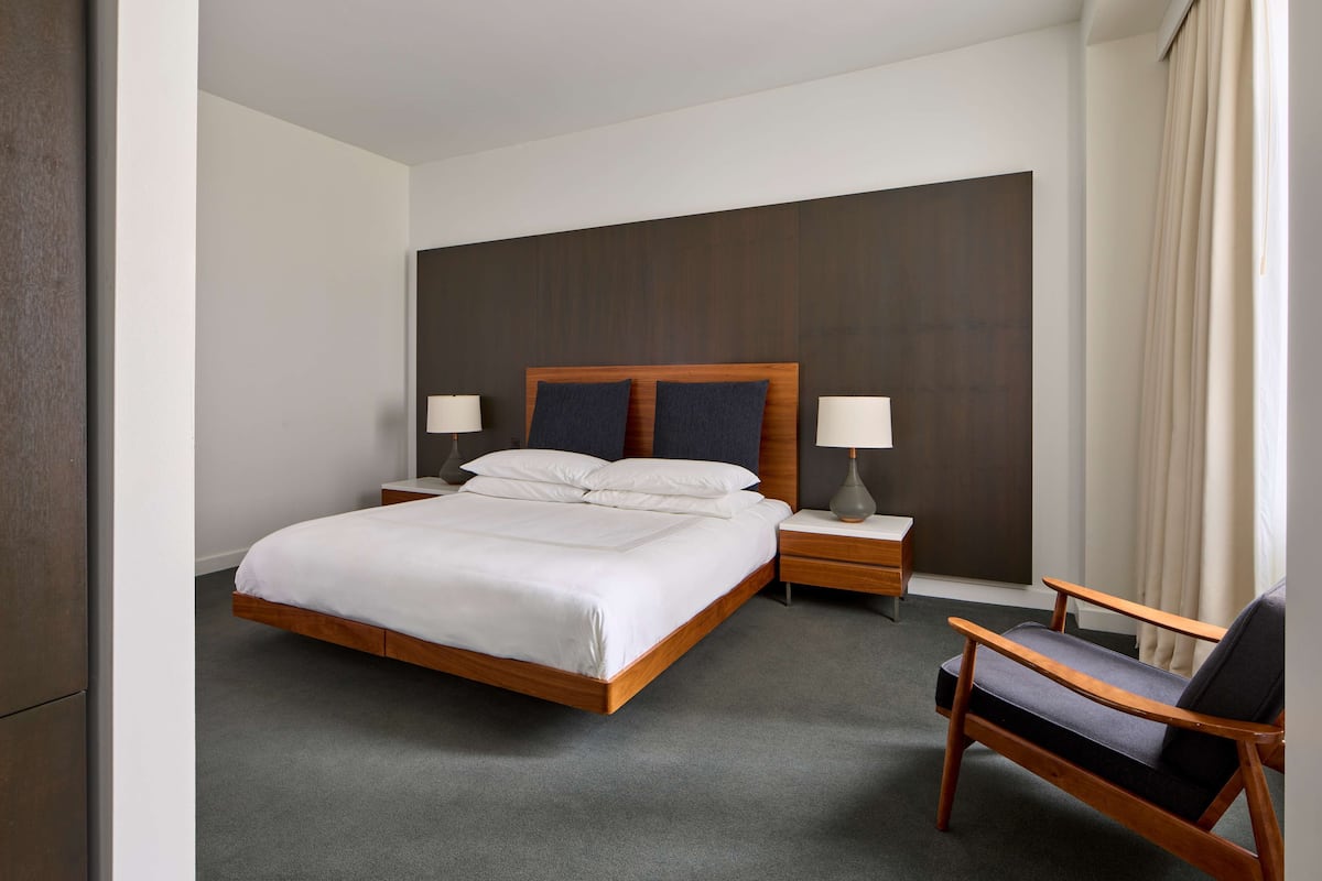 A hotel room with a large bed, two bedside tables with lamps, a wooden chair, and a view of the outside through a window with curtains.
