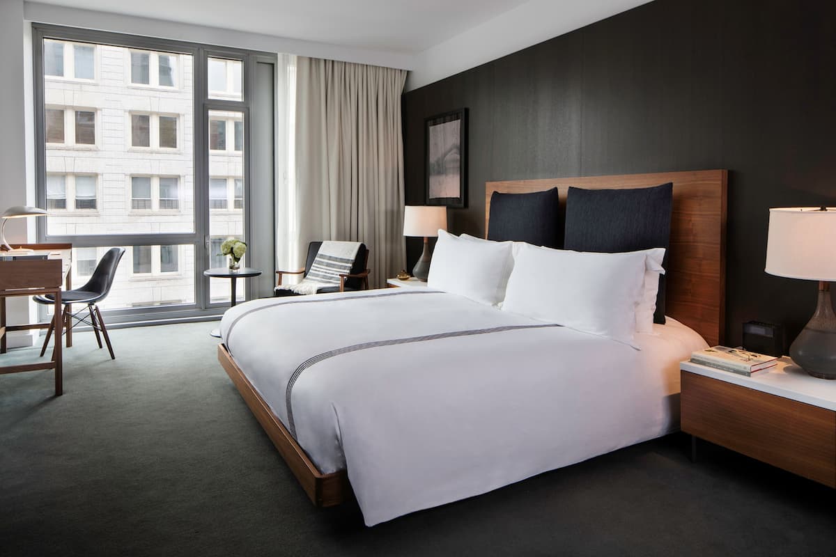 A hotel room with a large bed, a desk, a chair, a nightstand, and a view of a building through a window.
