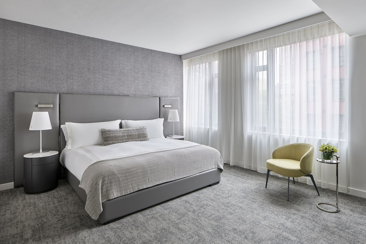 A modern hotel room with a large bed, two bedside tables with lamps, a chair, and a view of the city through sheer curtains.